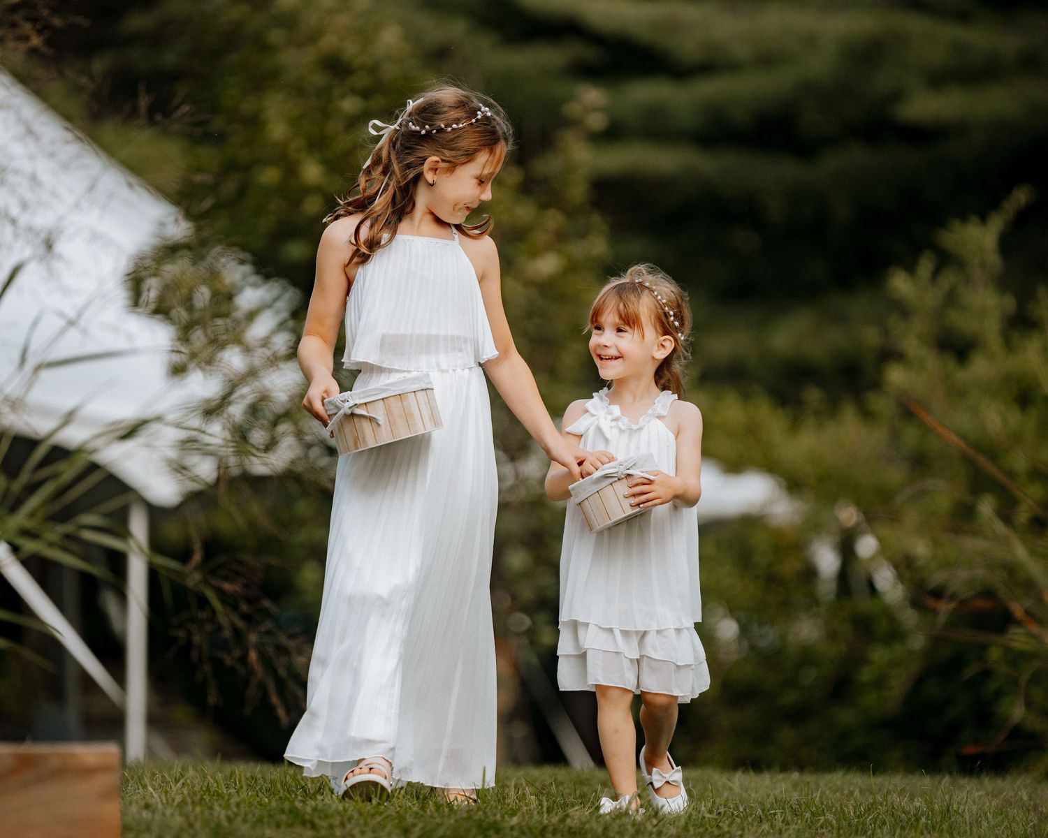 Two flower girls in white dresses walk on grass carrying baskets at an outdoor wedding ceremony.