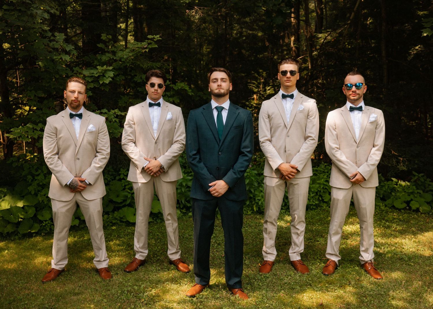 Groom in navy suit stands with four groomsmen in tan suits and bow ties outdoors in a wooded setting.