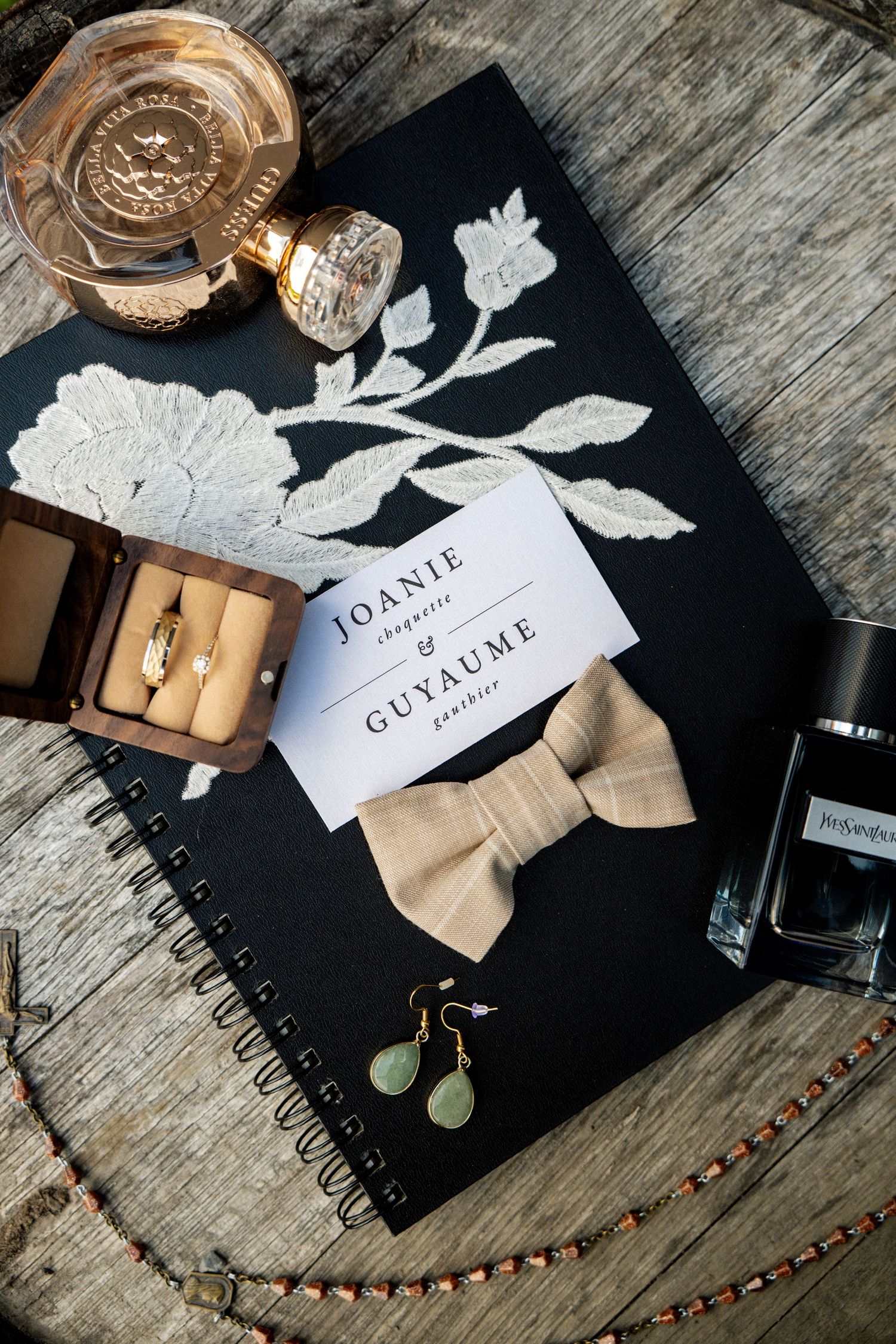 Elegant wedding flat lay featuring invitation cards for Joanie and Guyaume, a bow tie, rings, earrings, and perfume on dark background.