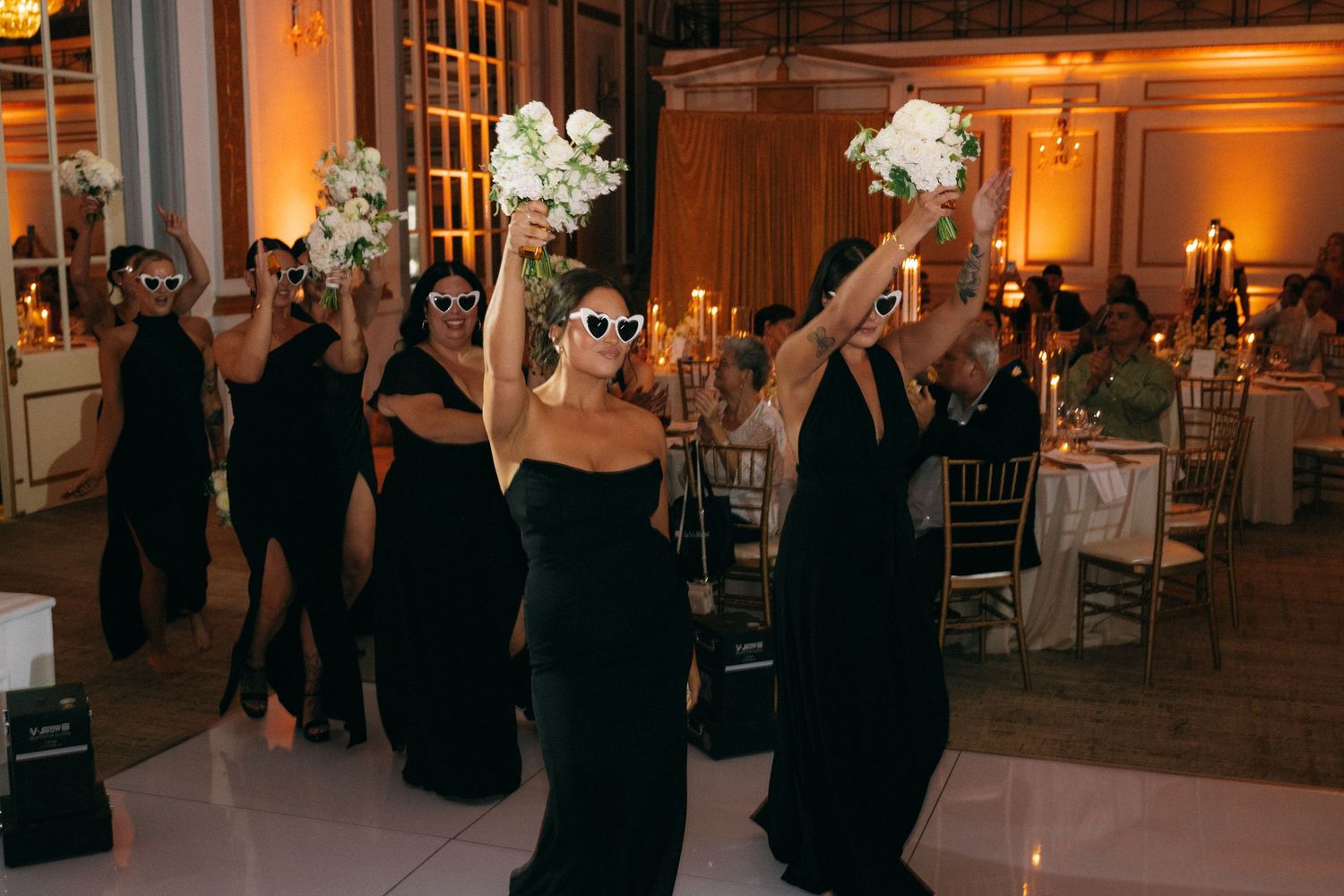 Bridesmaids in black dresses wearing sunglasses raising white bouquets on dance floor at wedding reception.