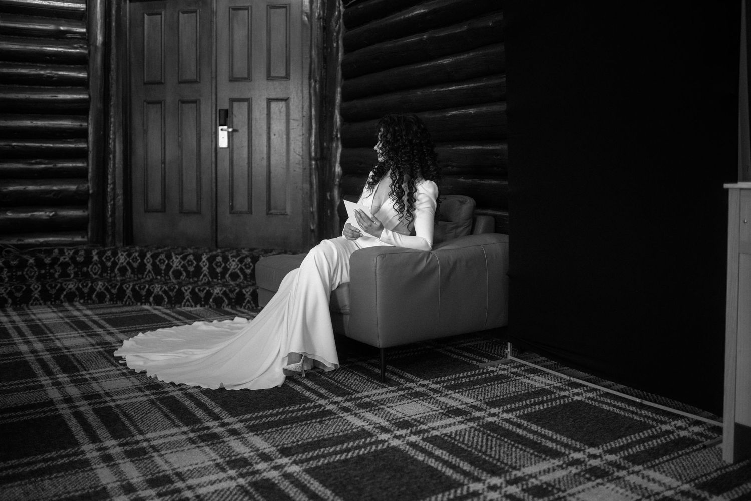 Bride in elegant white gown sitting alone on a chair in a moody black and white wedding photo.