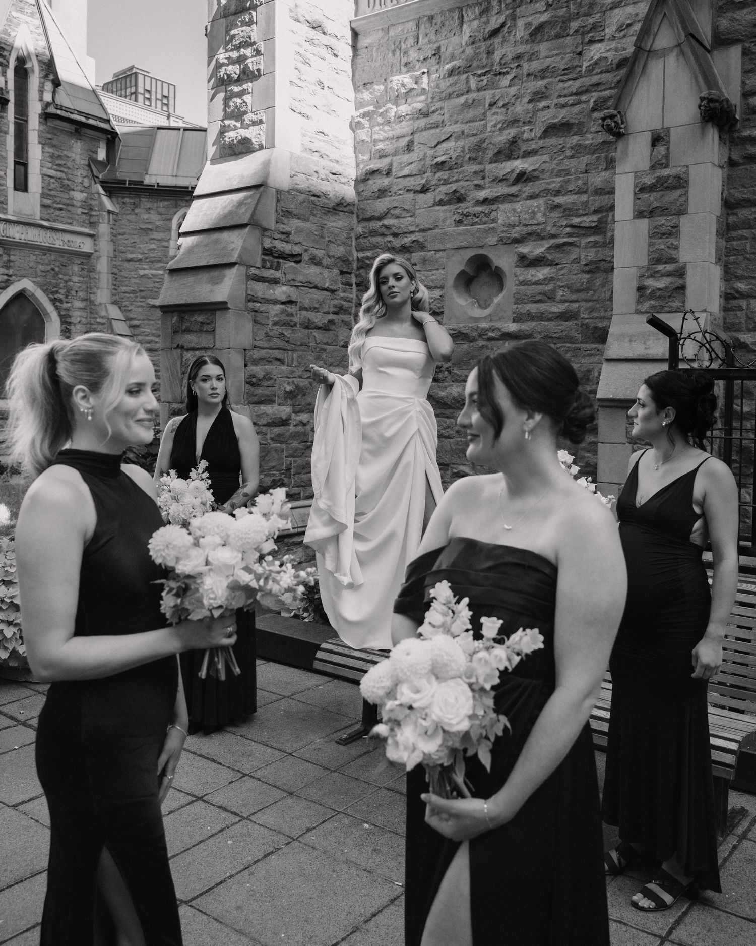 Bridesmaids in black gowns holding white bouquets chat outside a stone church while the bride stands behind them.