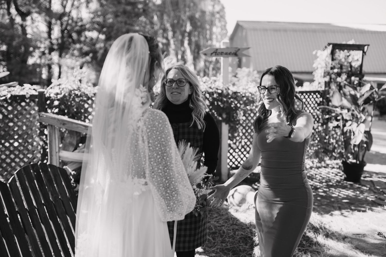 Bride in white wedding gown and veil greeting guests outdoors in black and white photo.