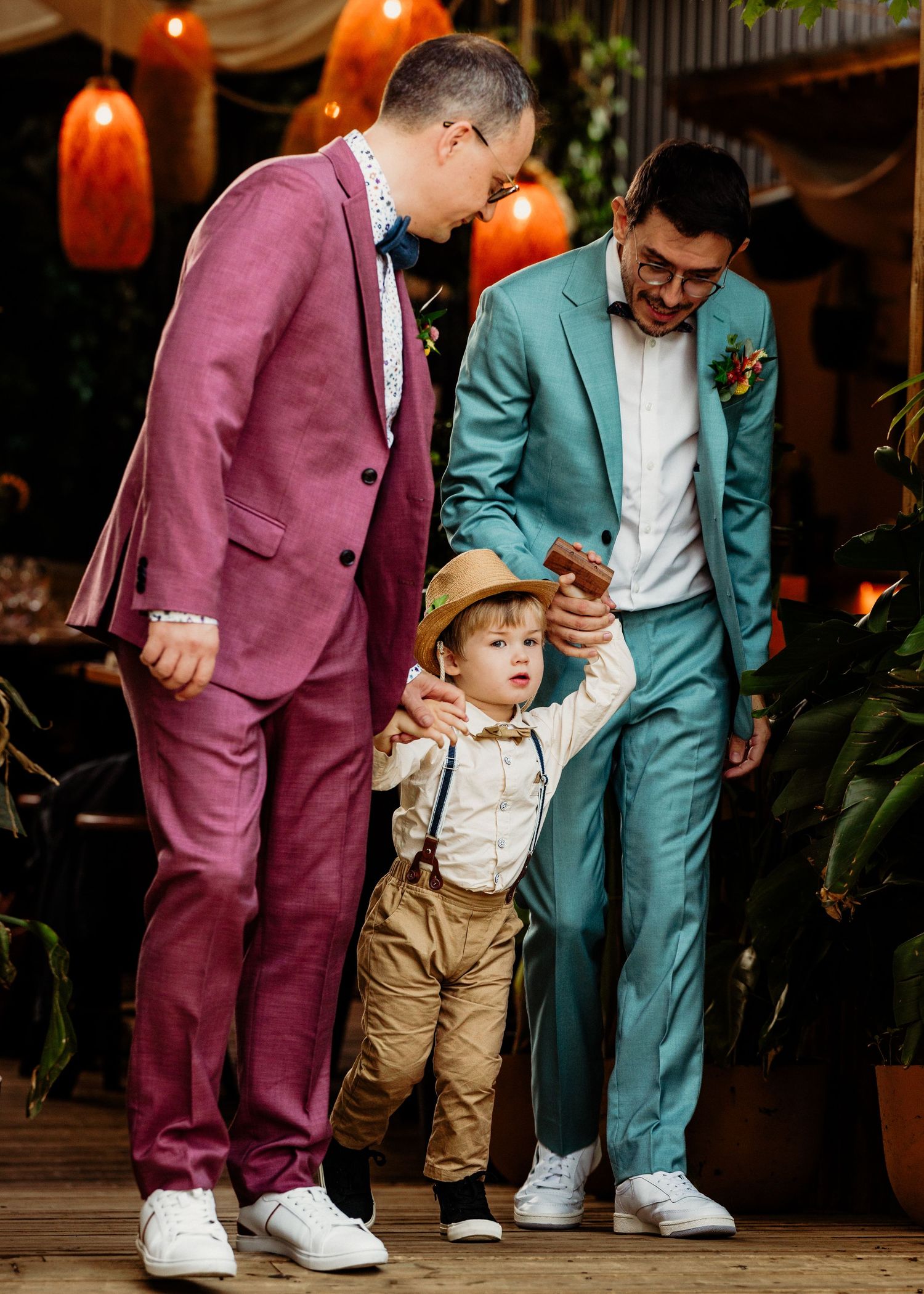 Two grooms in pink and teal suits help their young son walk at their wedding celebration.