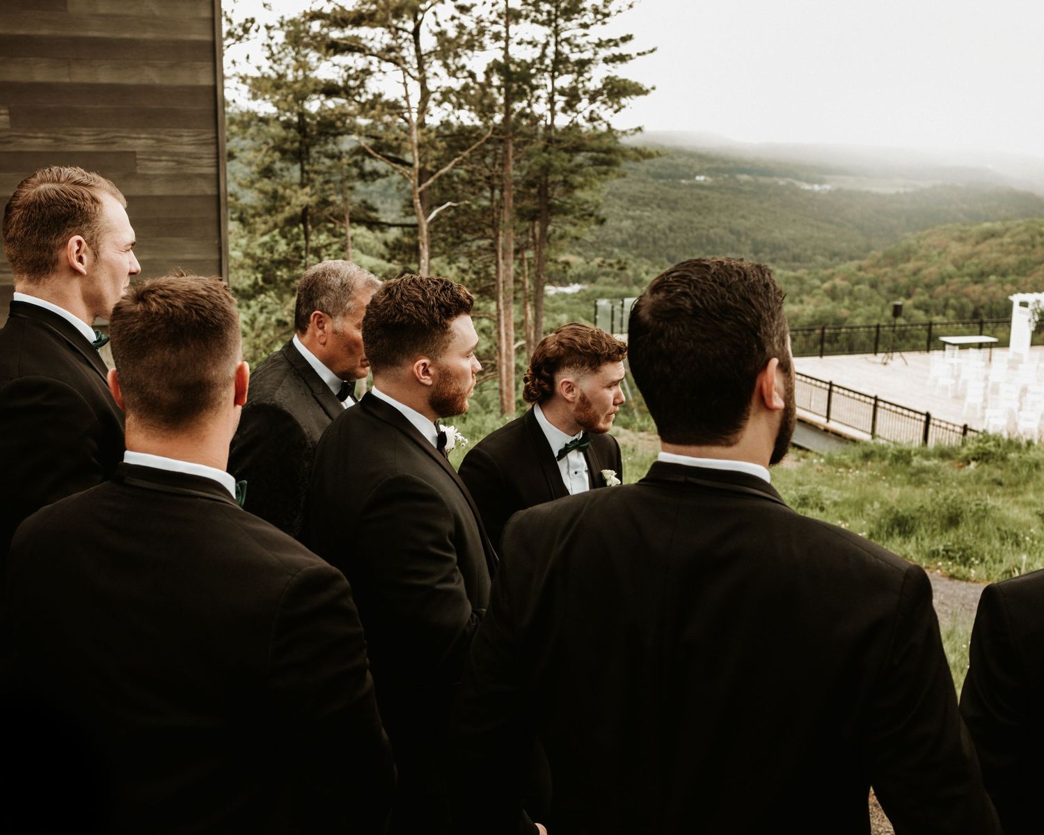 Groomsmen in black tuxedos gather outdoors at a scenic mountain wedding venue with lush green hills.
