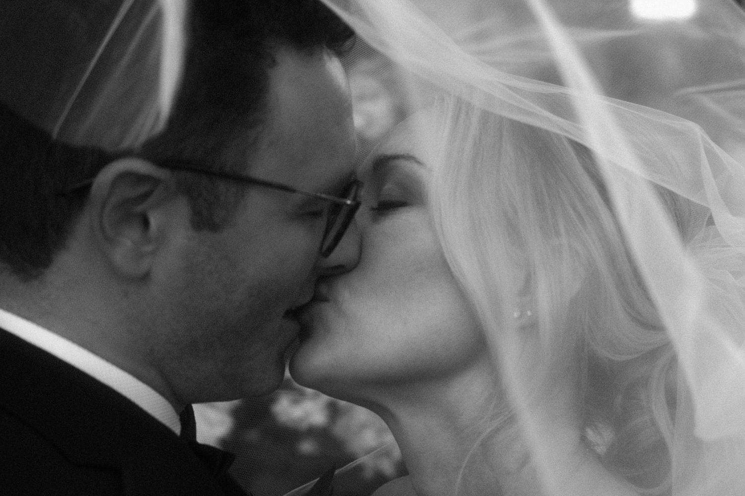 Romantic black and white close-up of bride and groom kissing under flowing wedding veil.