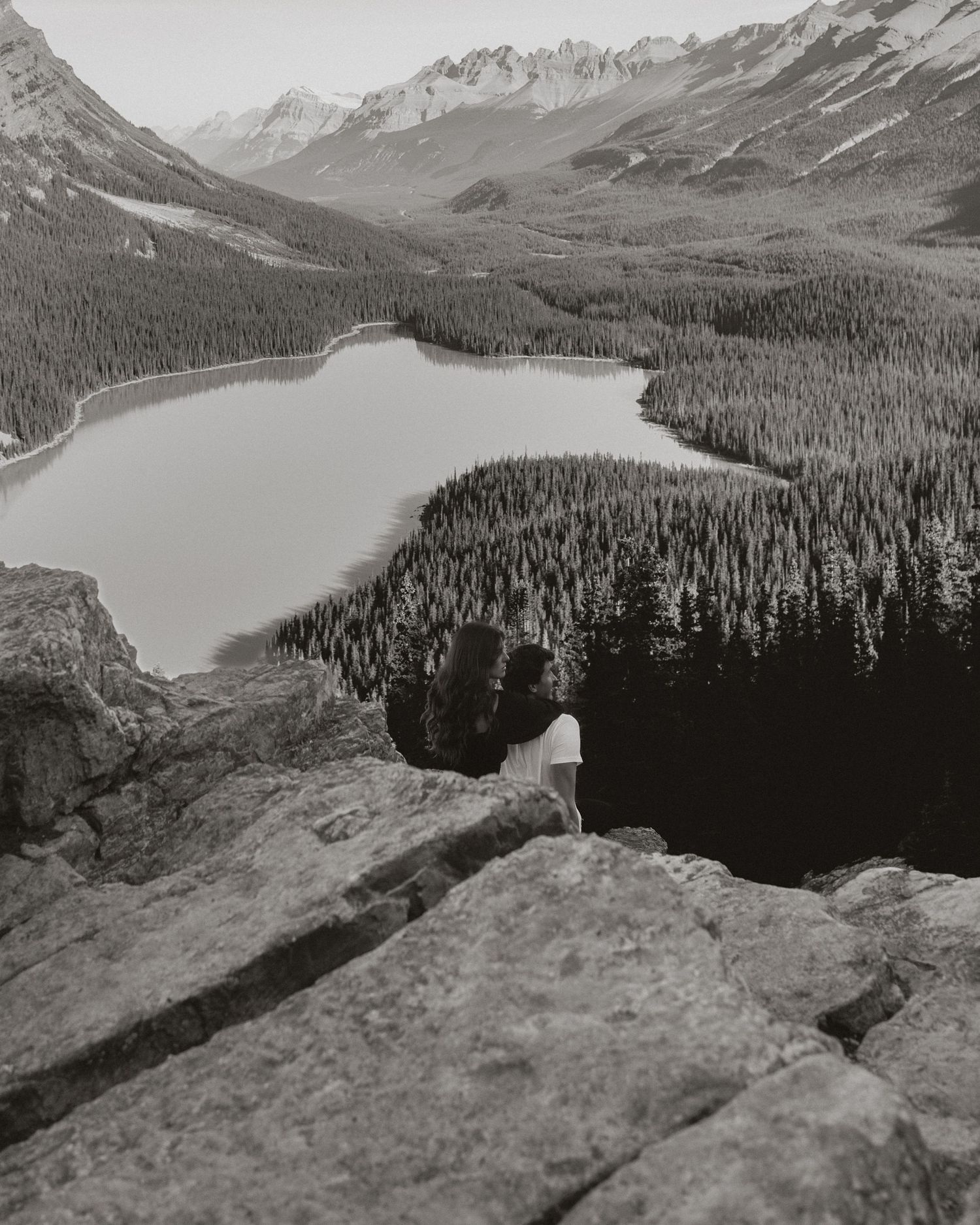 Hiker overlooks stunning glacial lake surrounded by pine forests and mountains in black and white landscape photography.