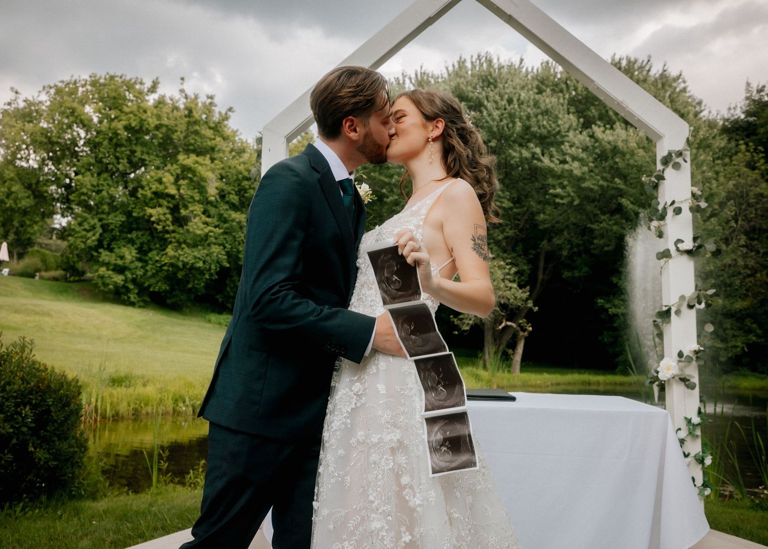 Newlywed couple sharing a first kiss at outdoor wedding altar, bride holding ultrasound photos, lush green garden backdrop.