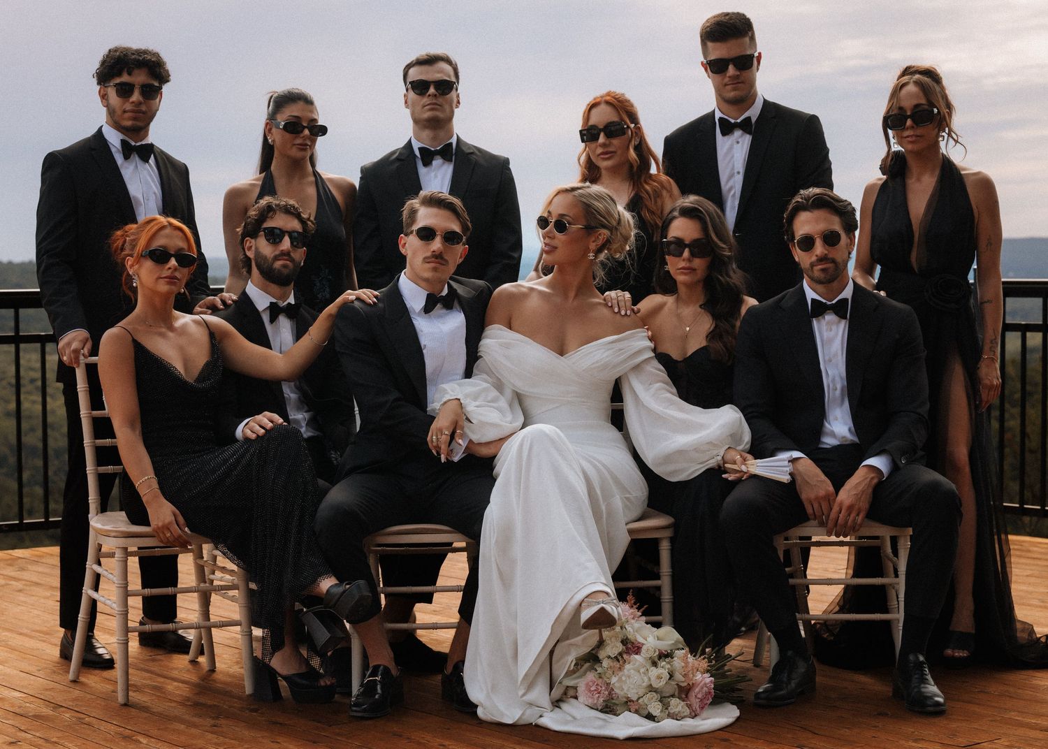 Wedding party poses together in elegant black formal attire with bride in white off-shoulder gown on scenic outdoor terrace.