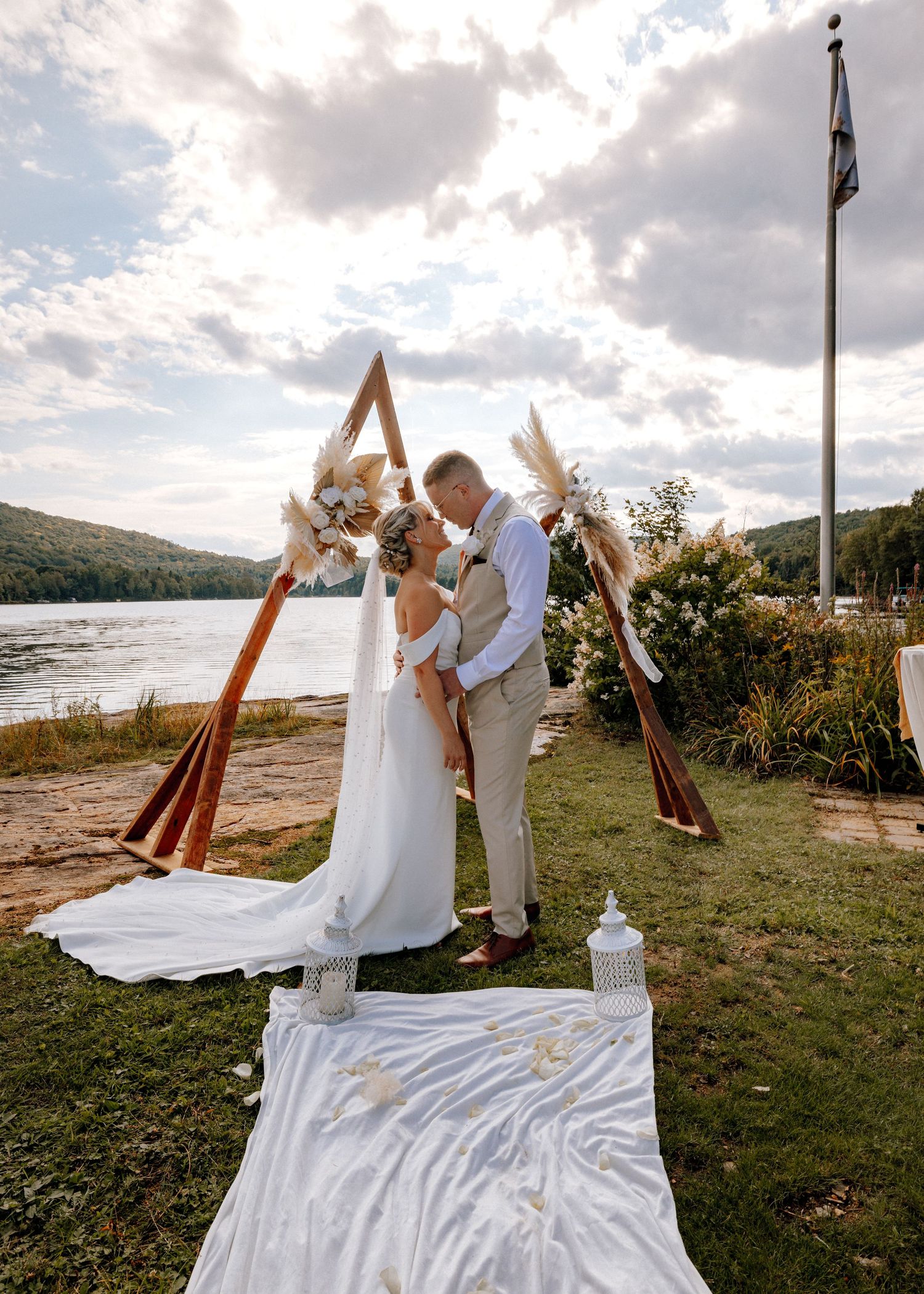 Bride and groom kissing under a boho triangular arch by a lakeside during their outdoor wedding ceremony.