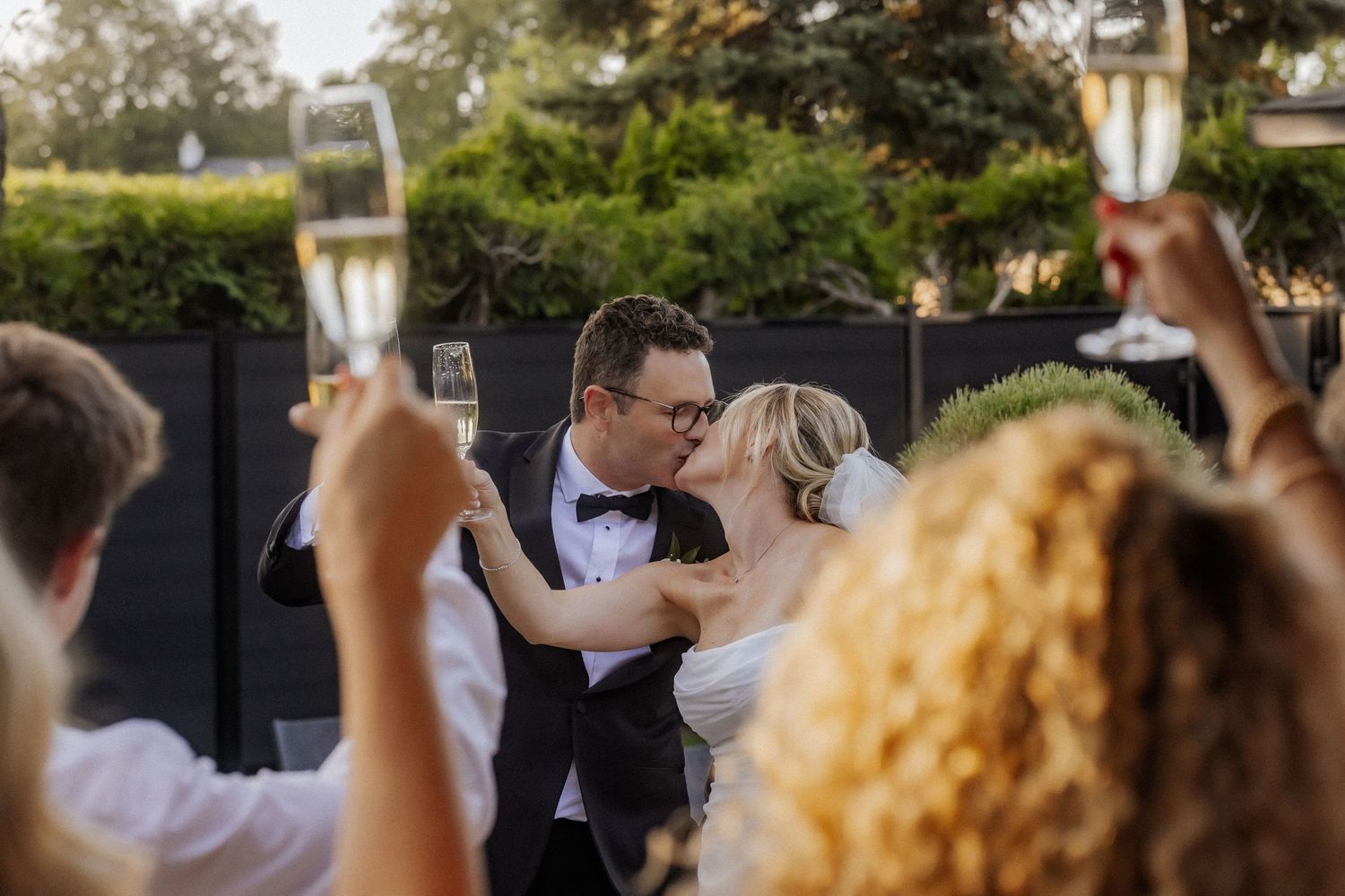 Bride and groom kiss as guests raise champagne glasses in celebratory wedding toast outdoors.
