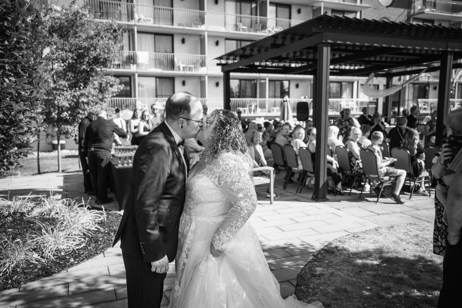 Black and white photo of a bride and groom sharing a kiss at an outdoor wedding ceremony with guests seated nearby.