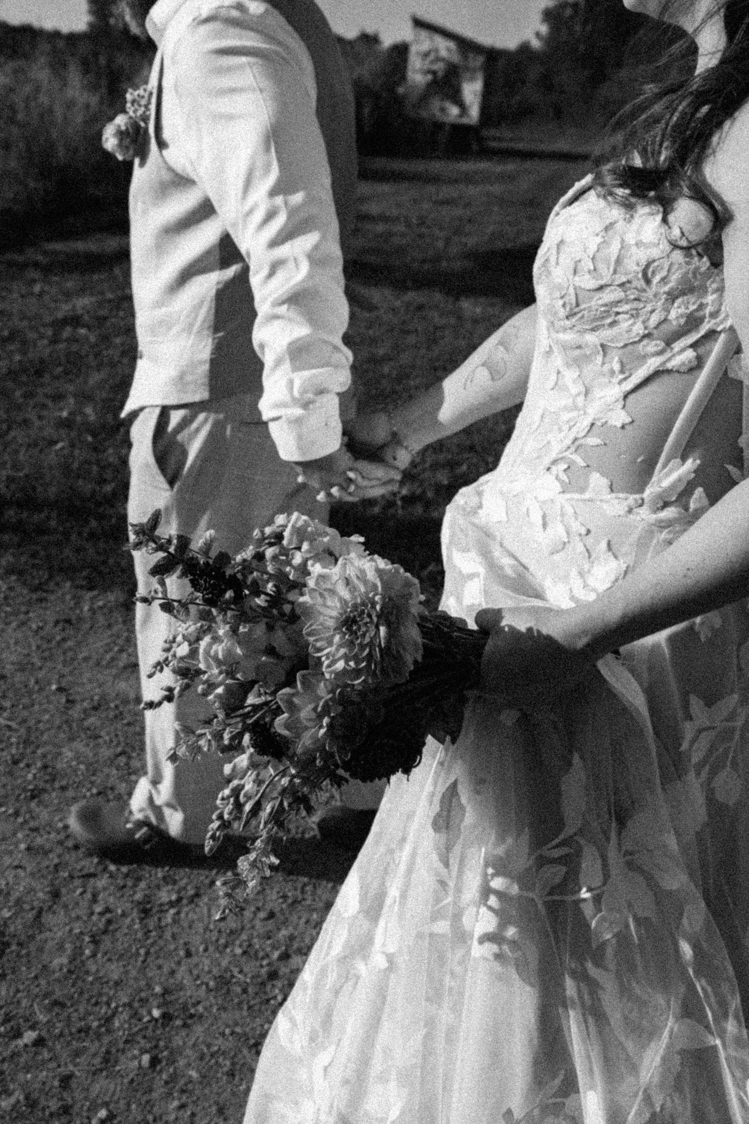 Black and white photo of bride holding floral bouquet walking with groom outdoors.