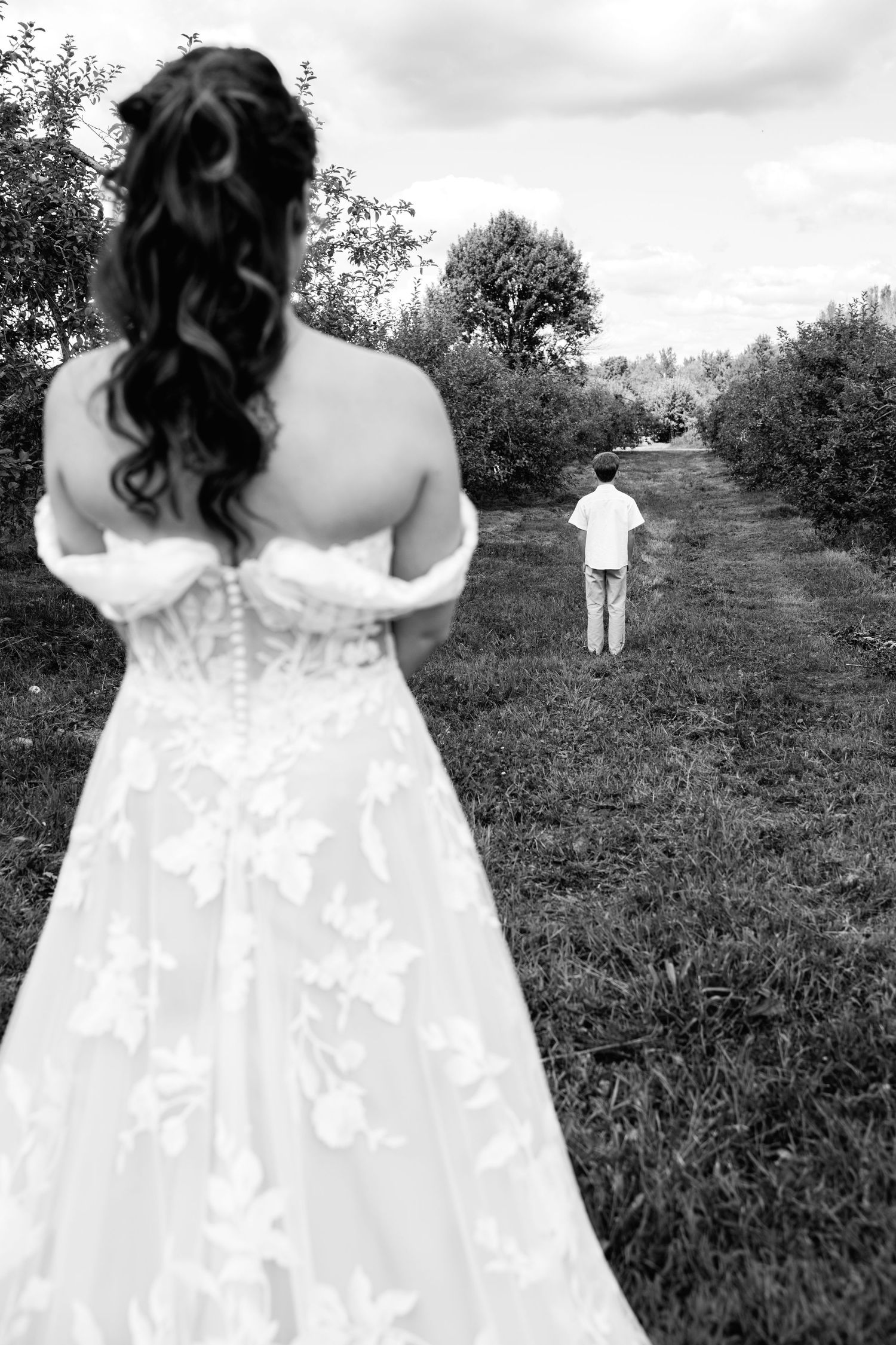 Black and white photo of a bride in a lace wedding gown viewed from behind, facing a child walking down a garden path.