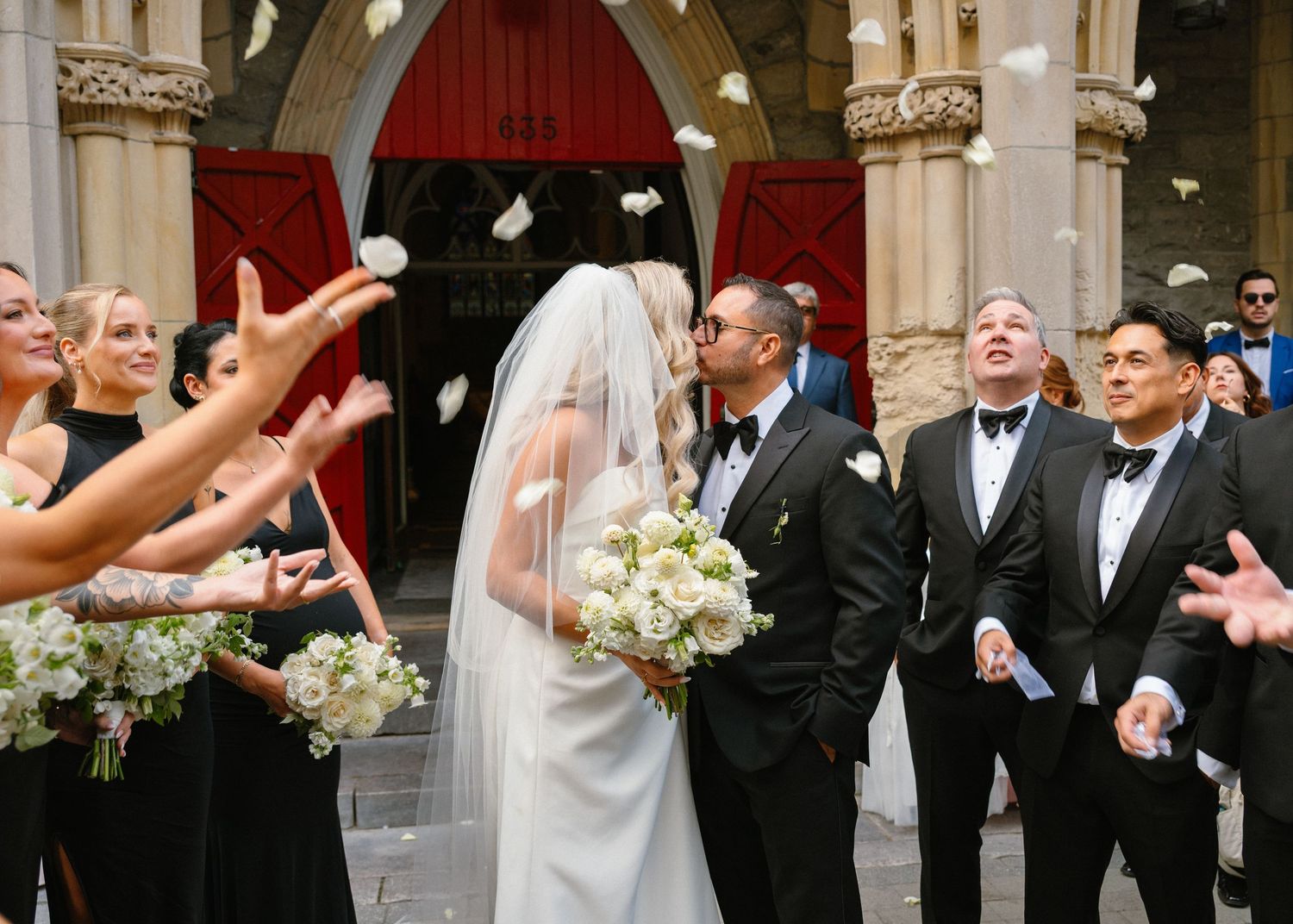 Bride and groom kiss outside church as wedding party tosses white flower petals in celebration.