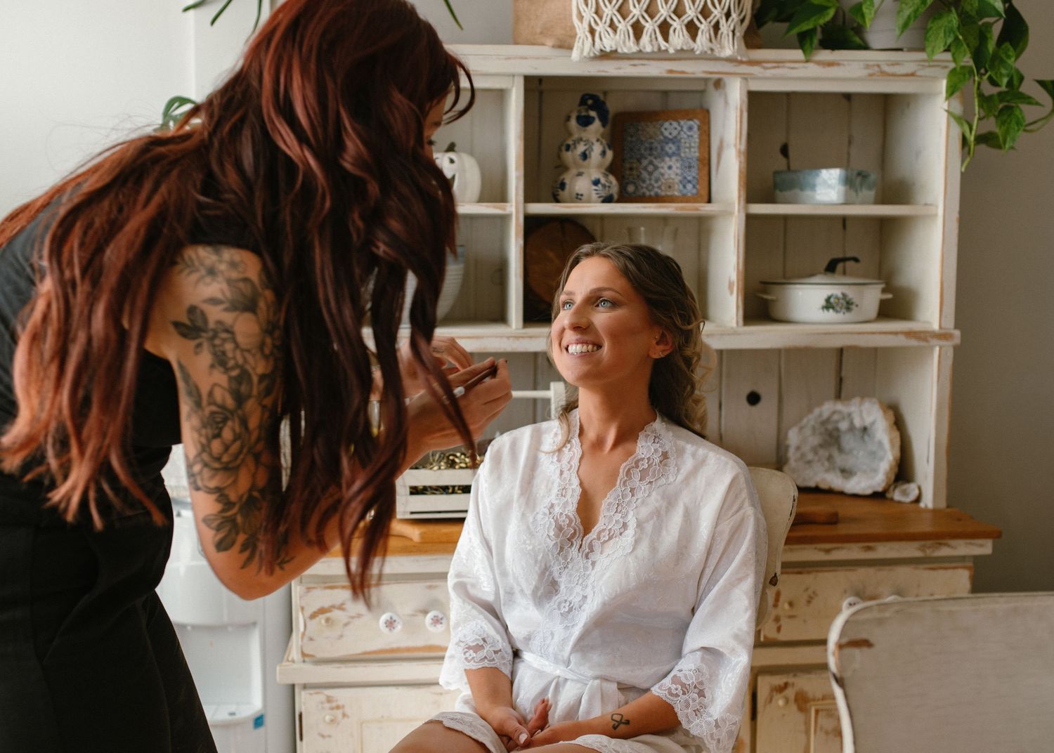 Tattooed hairstylist styling a smiling bride in a white robe inside a vintage-decorated room.