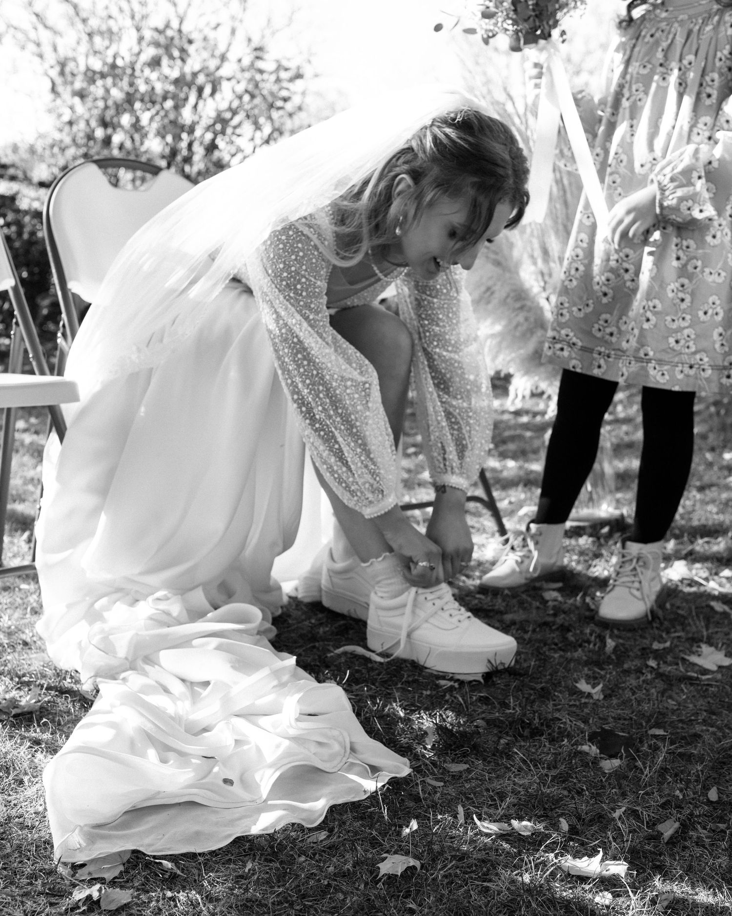 Bride in wedding dress and veil tying sneakers on grass at outdoor wedding in black and white.