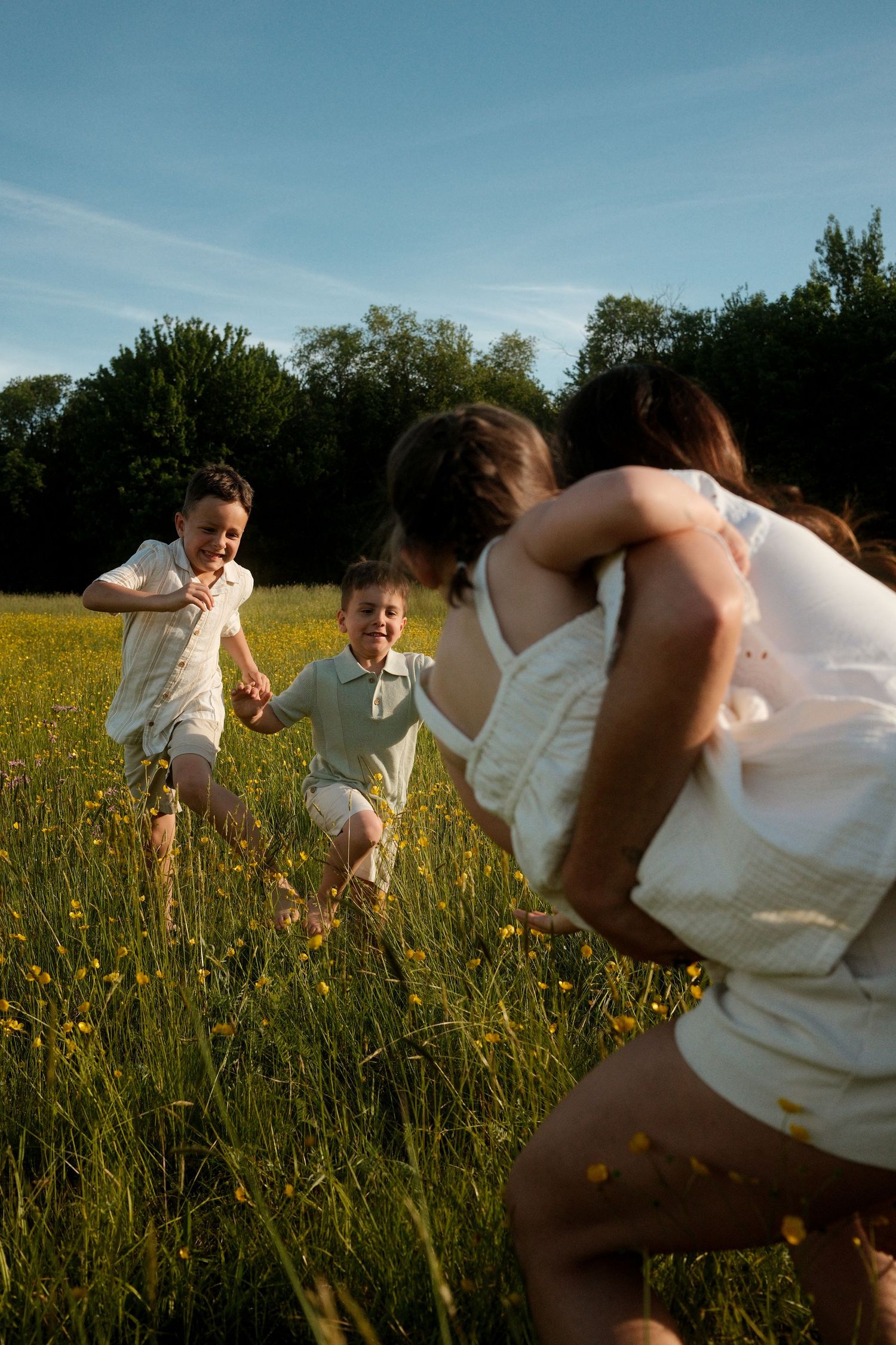 Two children joyfully run toward a woman in a sunny green meadow with yellow wildflowers.