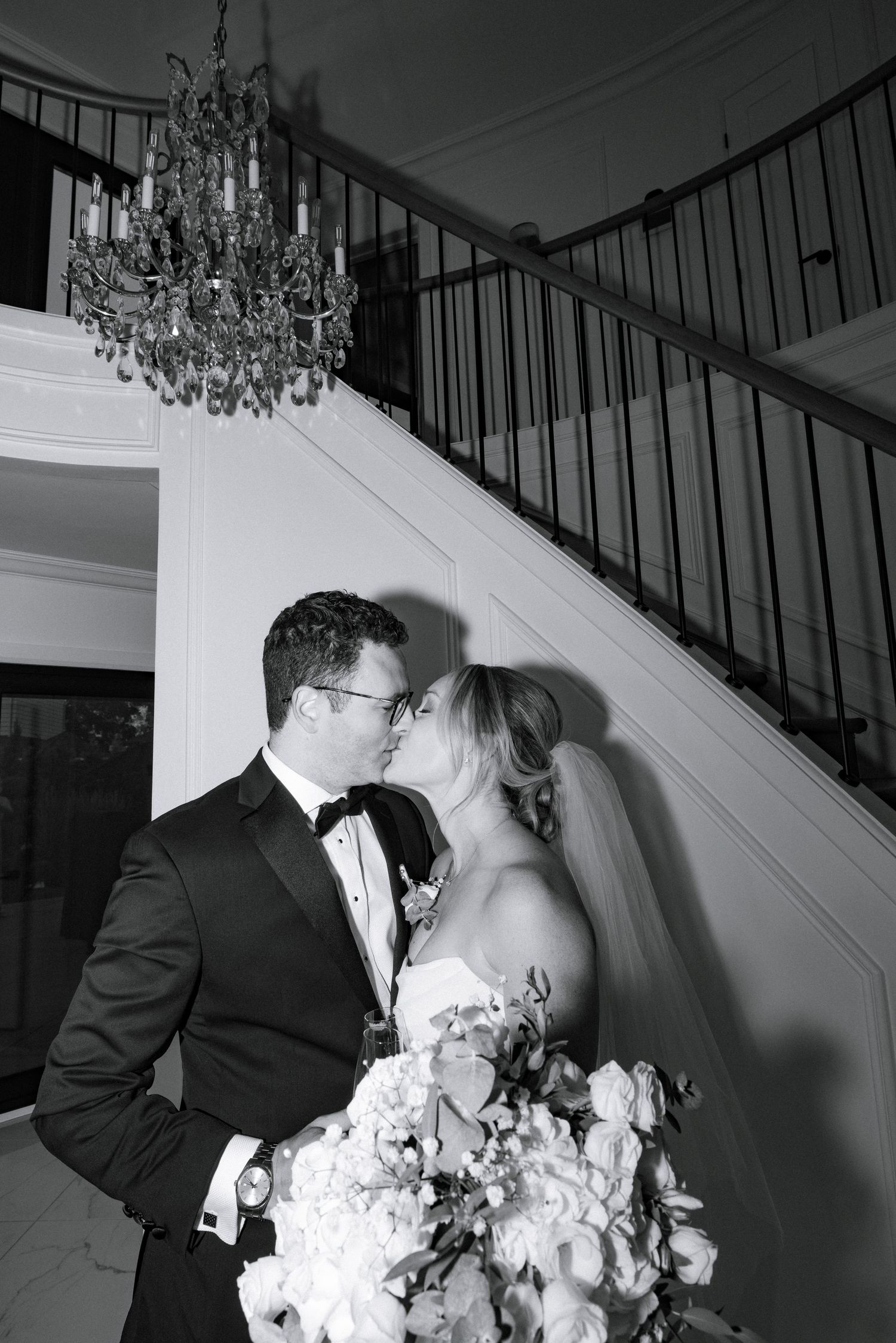Black and white photo of a bride and groom sharing a kiss near an elegant staircase with a crystal chandelier.