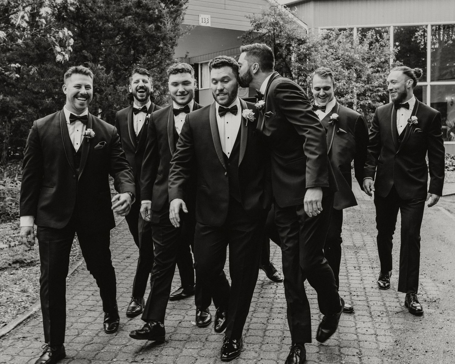 Groomsmen in black tuxedos laughing and walking together outdoors in a black and white wedding photo.