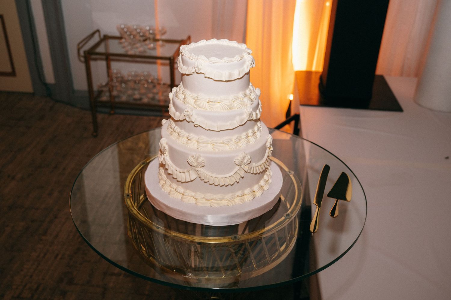Elegant four-tier white wedding cake with pearl and ruffle details displayed on glass table.