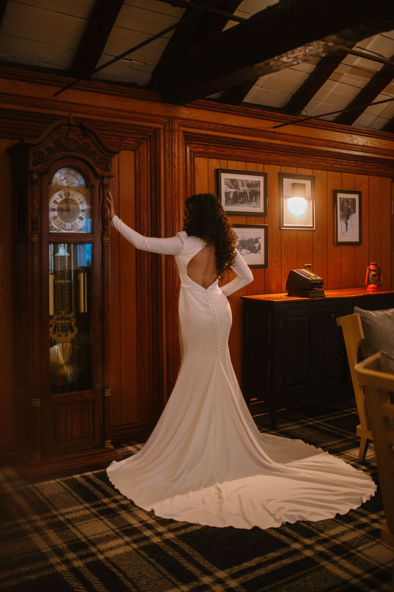 Bride in elegant backless white gown with train standing beside antique grandfather clock in wood-paneled room.