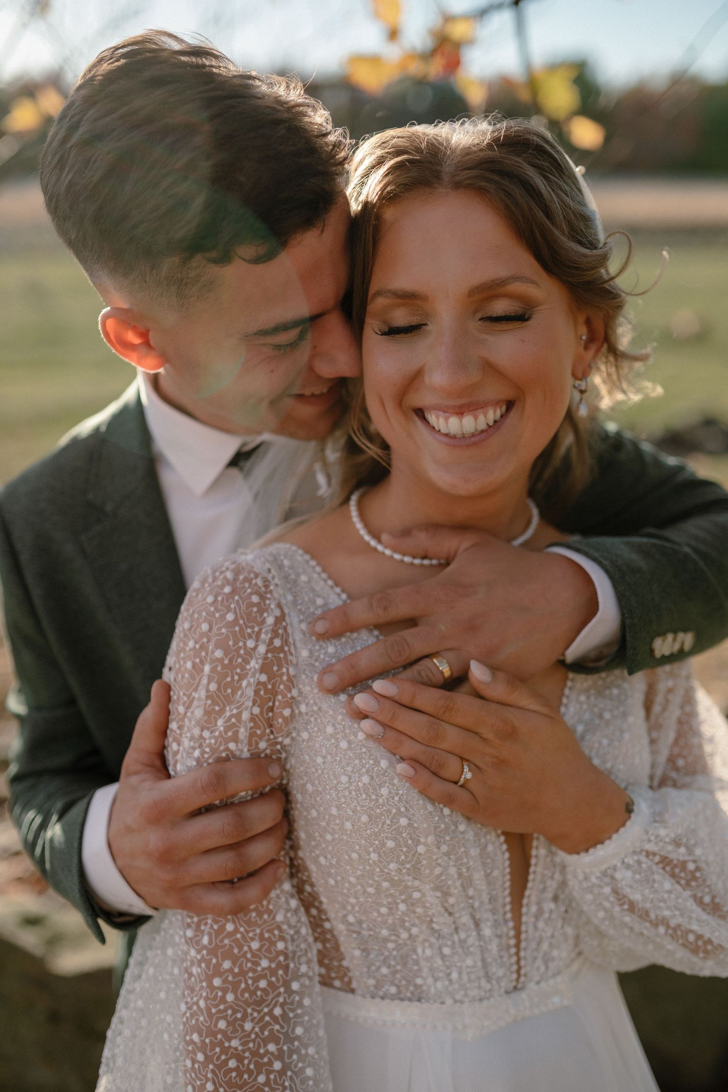 Bride and groom embrace at golden hour, she smiles in a beaded white dress as he holds her in a green suit.