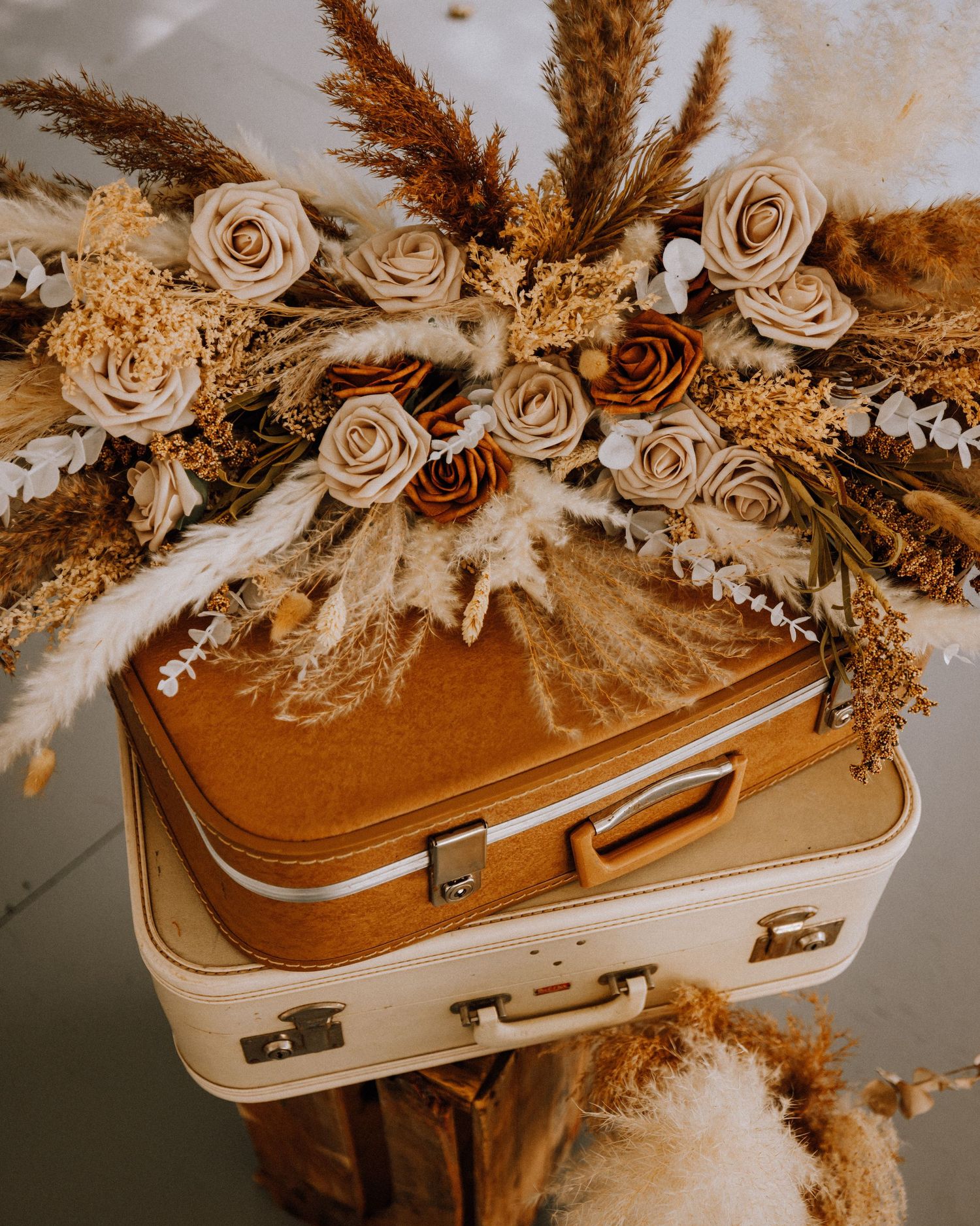 Vintage stacked suitcases topped with boho dried floral arrangement featuring pampas grass and cream roses.