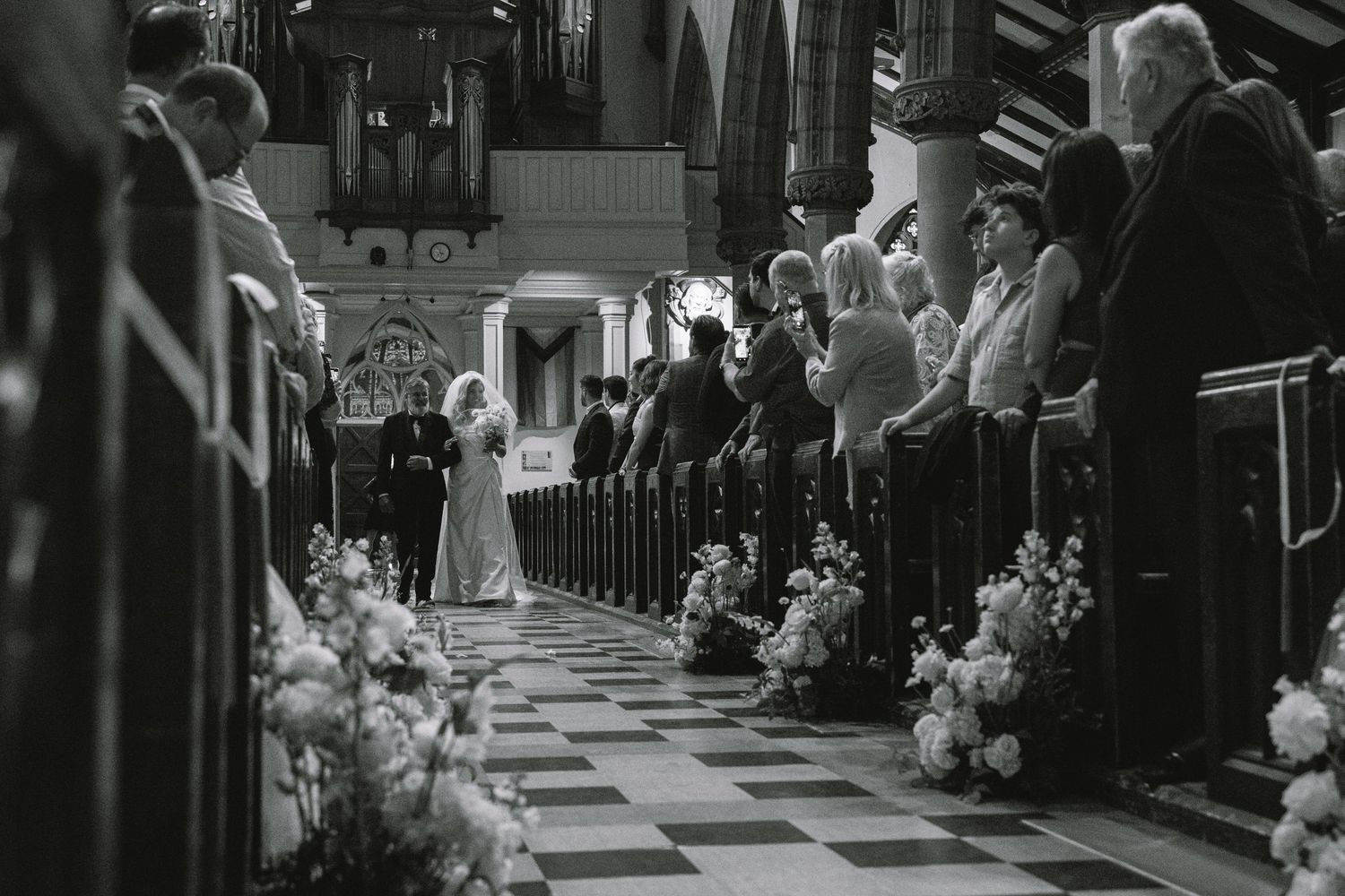 Black and white photo of bride walking down flower-lined aisle of traditional church with guests standing.
