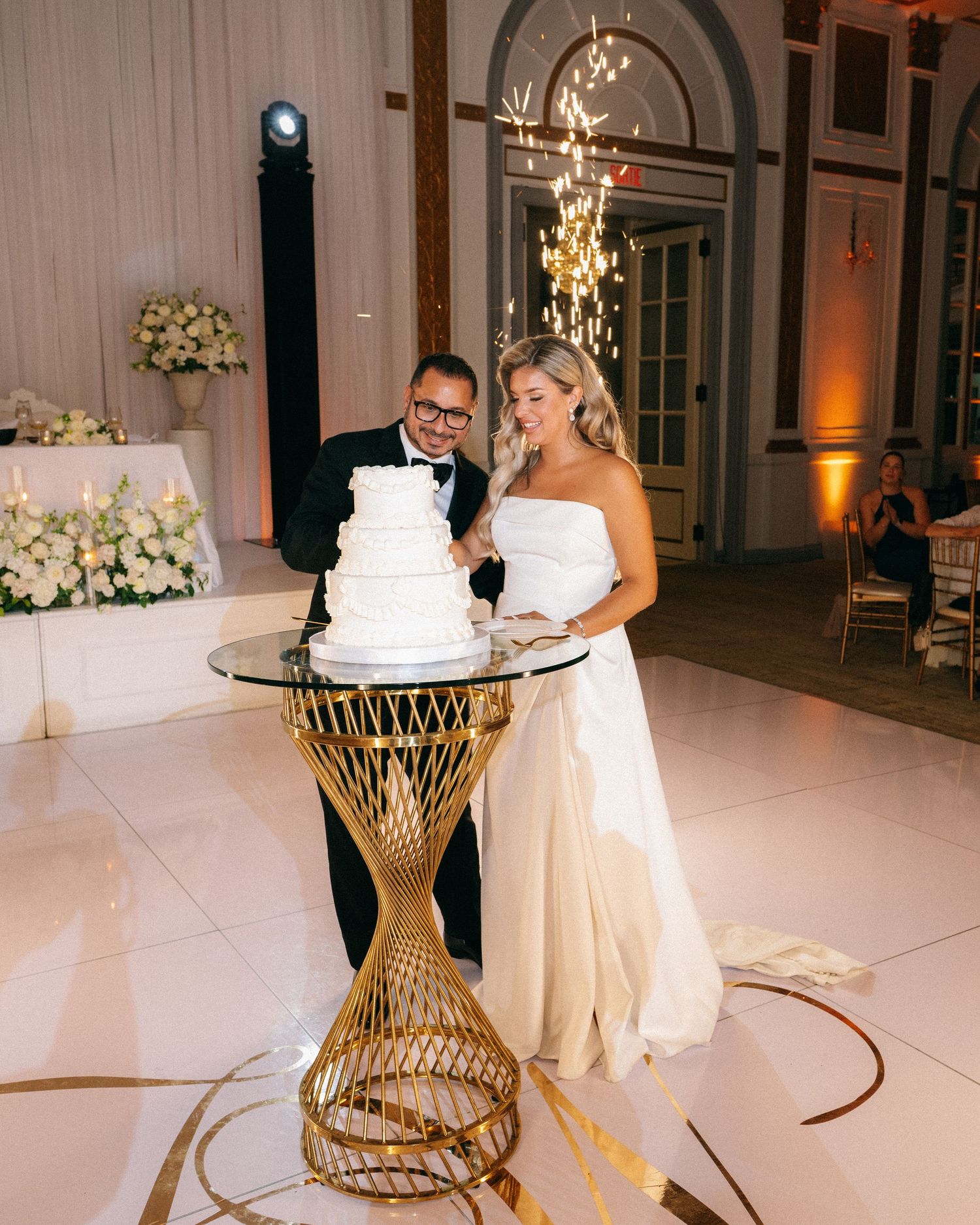 Bride and groom cutting tiered white wedding cake on gold geometric stand at elegant reception.