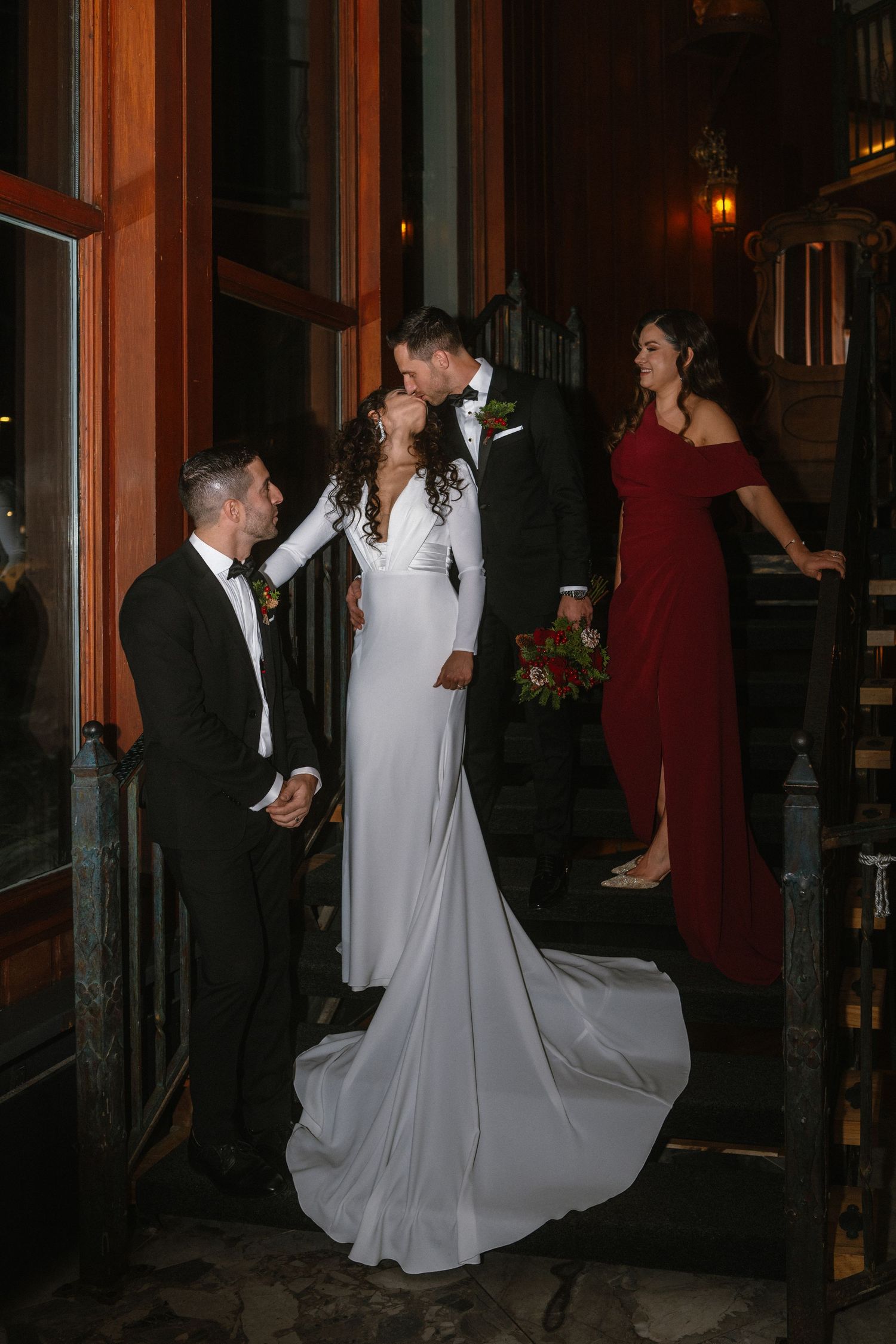 Bride and groom share a kiss on steps while groomsman and bridesmaid in red dress stand nearby at night.