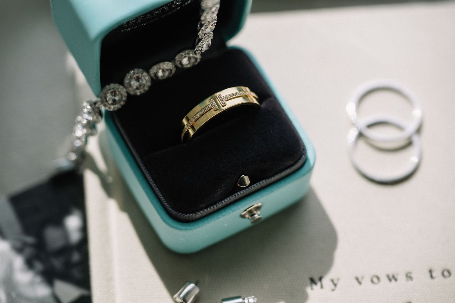 Gold Tiffany & Co. band ring in teal box with diamond bracelet and silver rings on vow book.