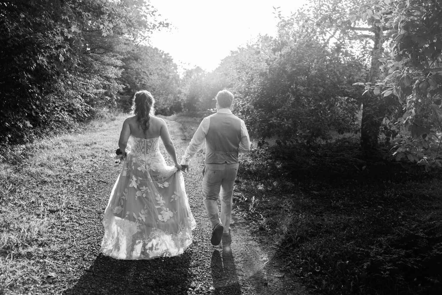 Bride and groom walk hand in hand down a sunlit garden path in romantic black and white wedding photo.