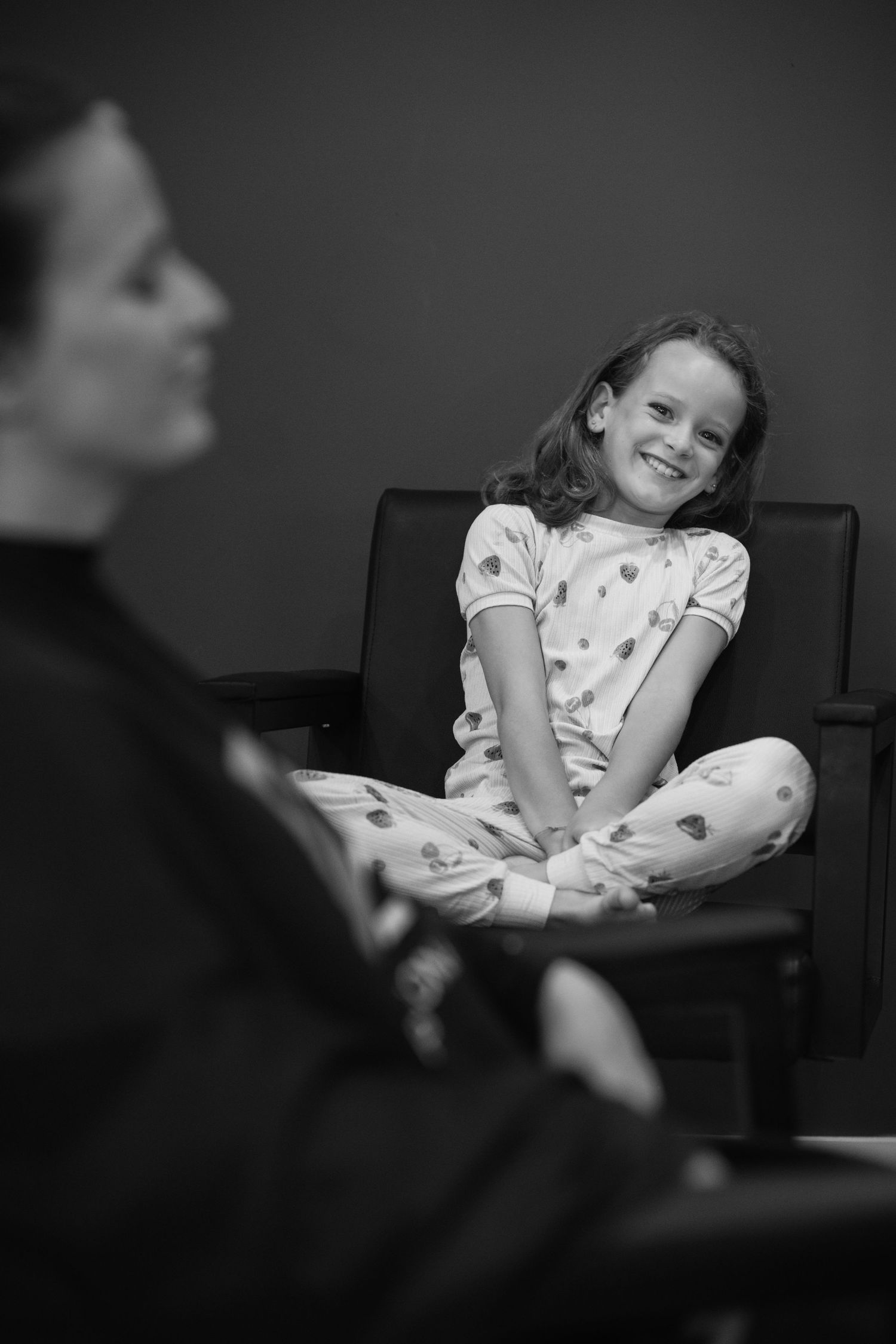 Black and white photo of a smiling girl in pajamas sitting cross-legged on a chair facing an adult.