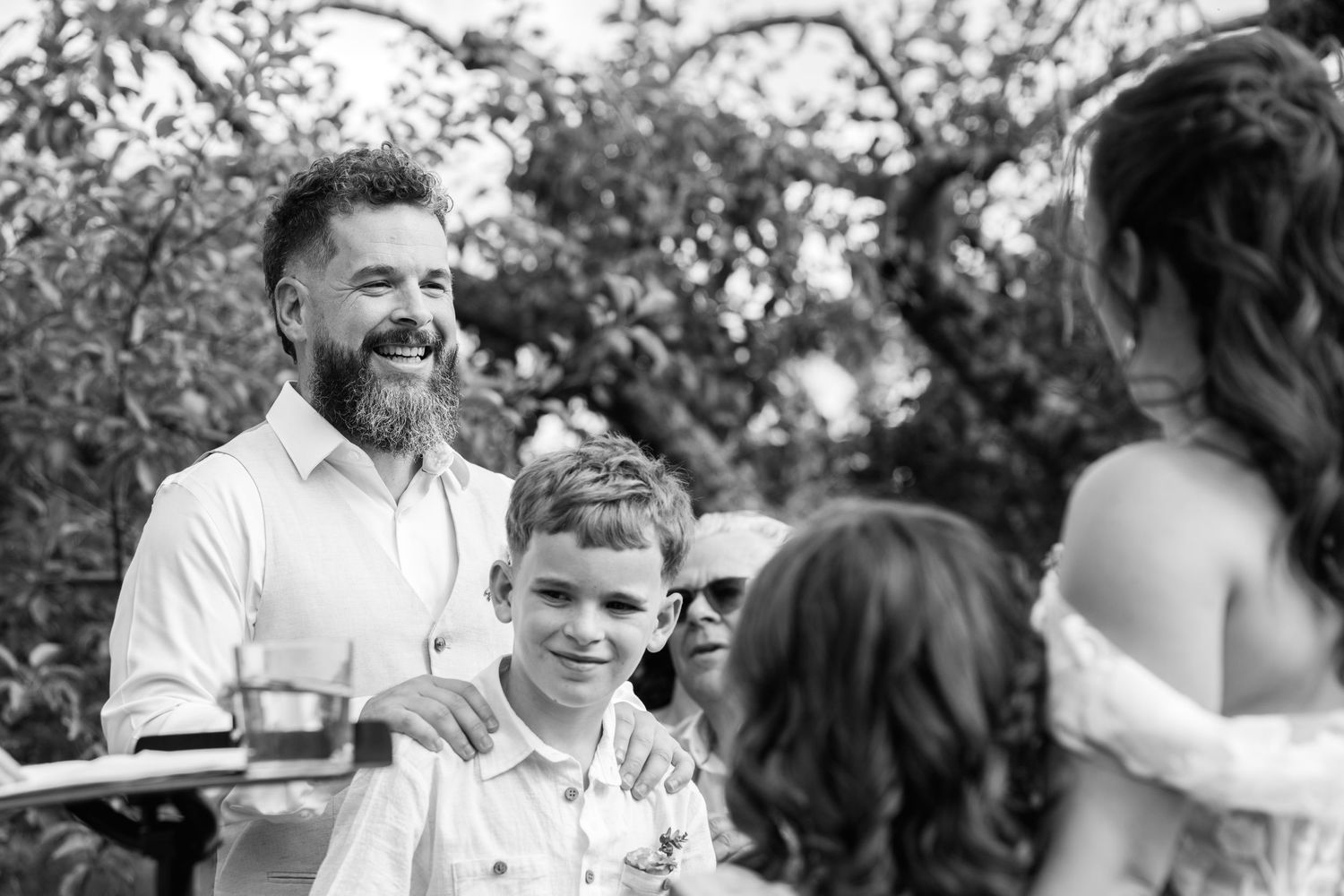 Bearded groom smiling with young boy at outdoor wedding ceremony in black and white.