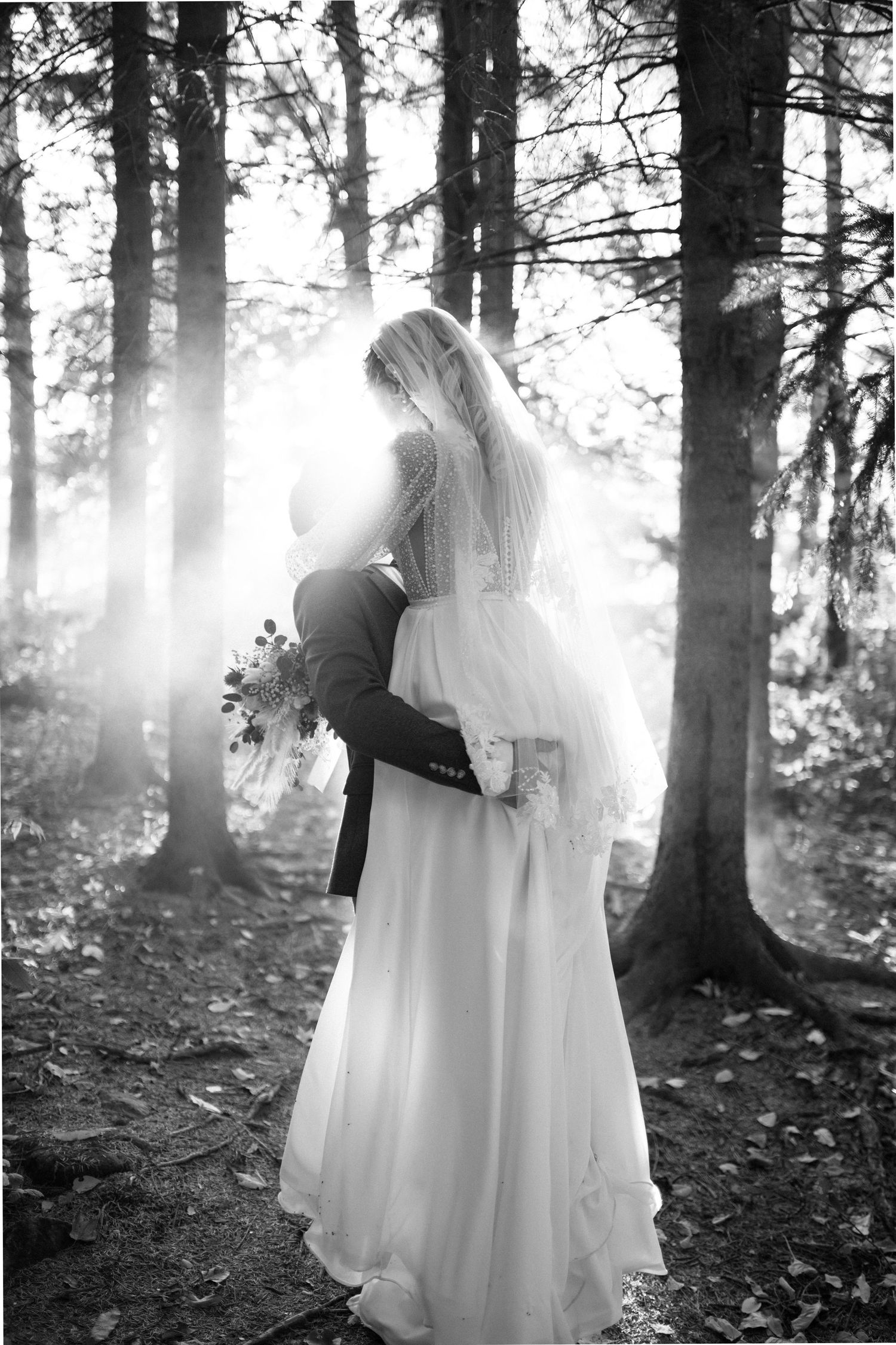 Black and white photo of groom lifting bride in flowing gown among sunlit forest trees.