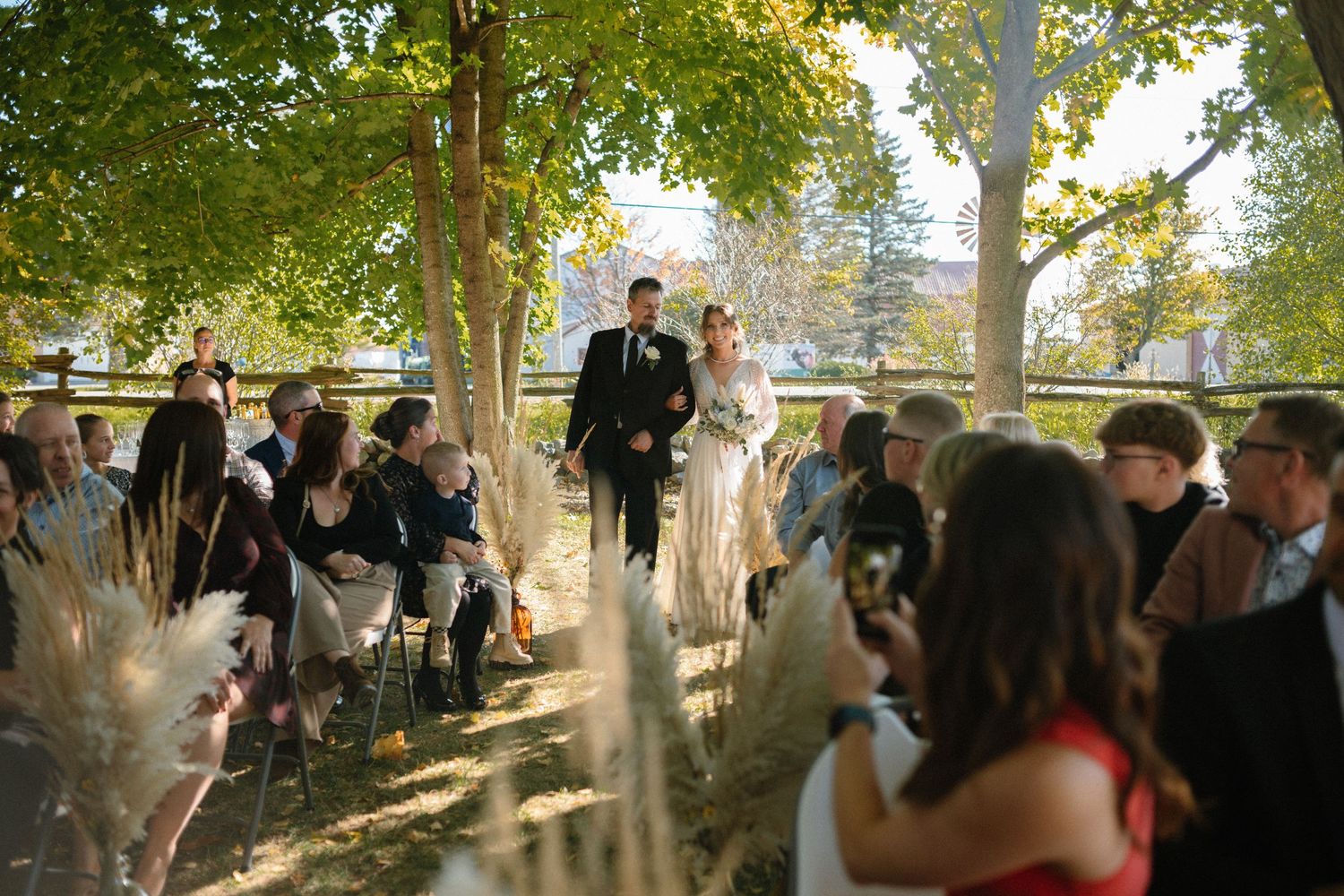 Bride and groom walk down outdoor aisle surrounded by guests and pampas grass decorations.