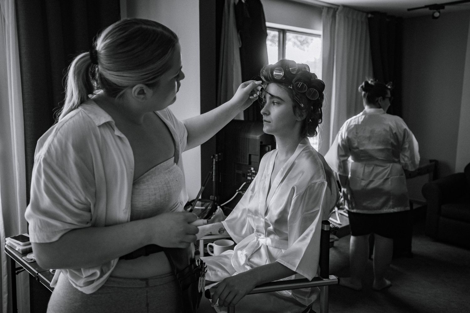 Bride getting hair and makeup done by stylists in a white robe before her wedding ceremony.