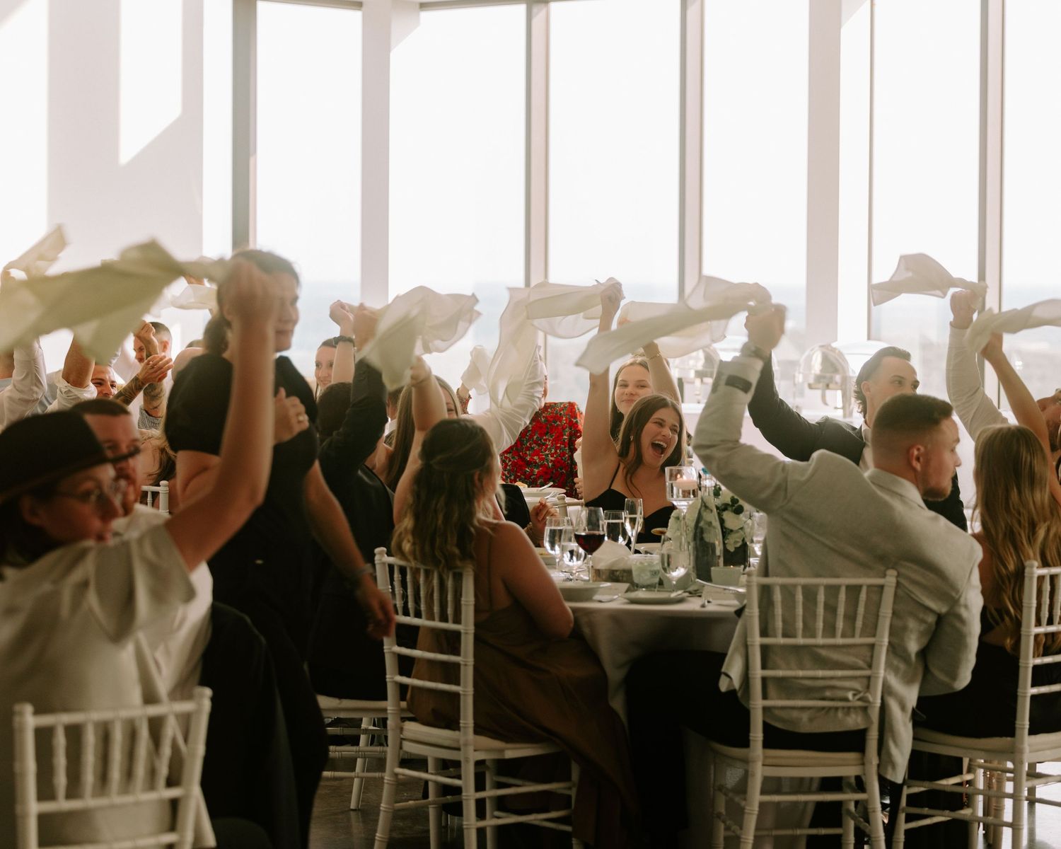 Wedding reception guests waving white napkins in elegant venue with large windows and white chiavari chairs.