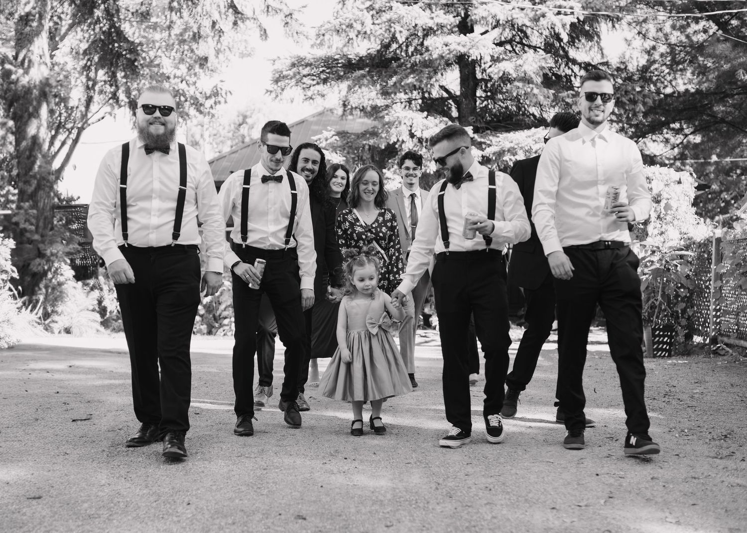 Black and white photo of groomsmen in suspenders and bow ties walking with flower girl.