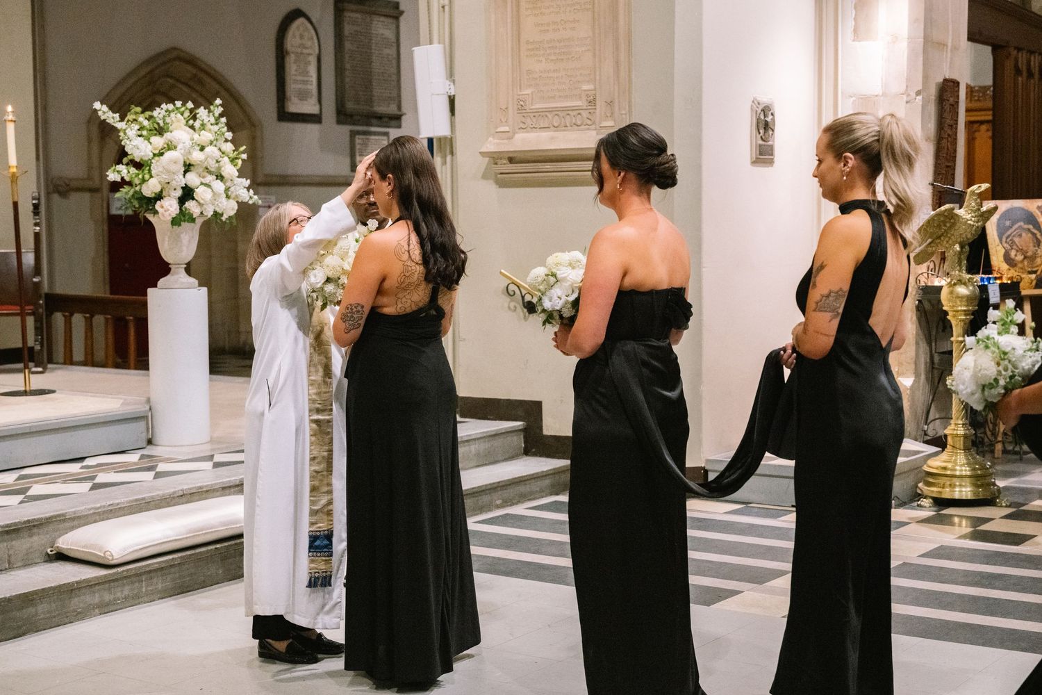 Bridesmaids in black strapless gowns holding white bouquets inside a church before the ceremony.