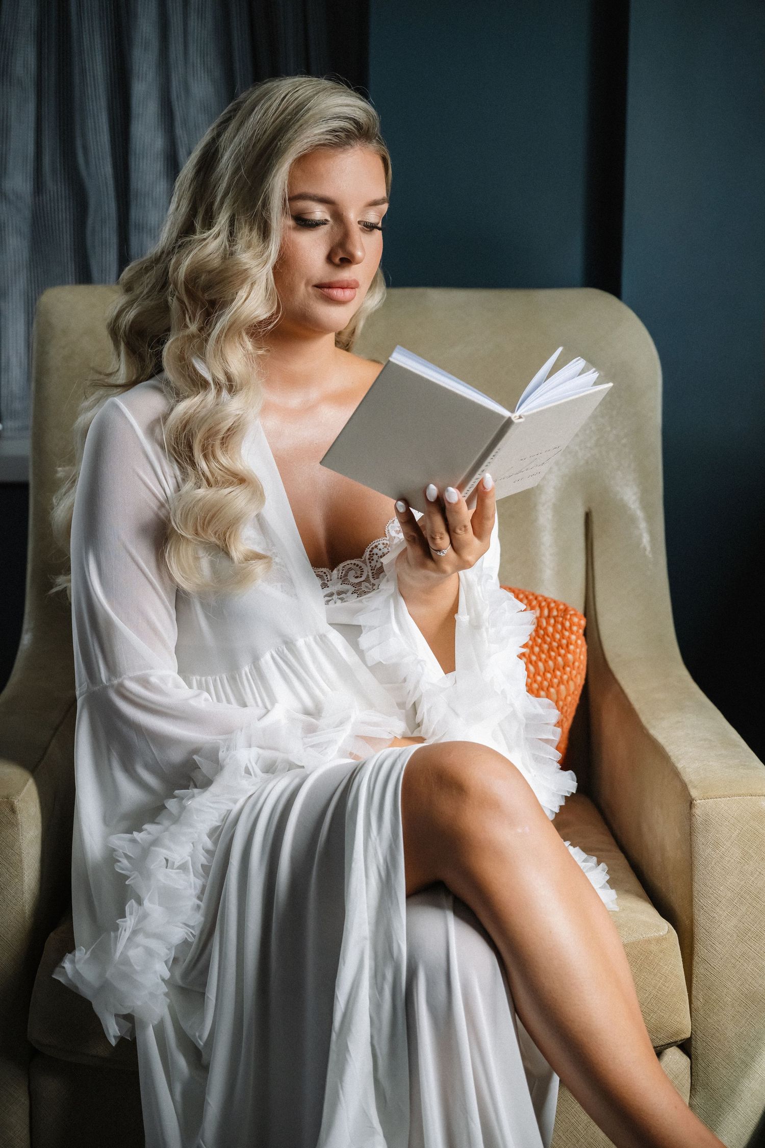 Elegant bride in white lace robe reading a book in an armchair before her wedding.