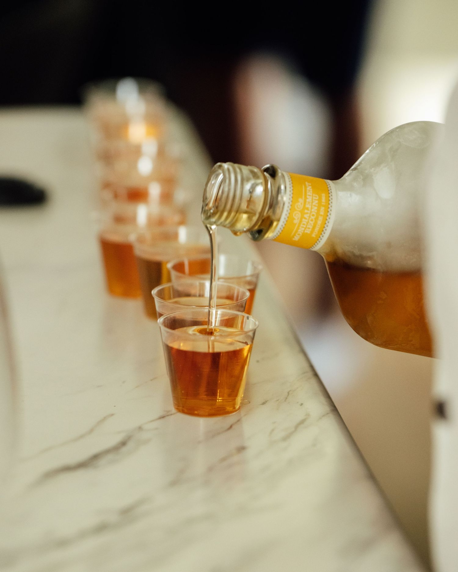 Amber liquid being poured from a bottle into small plastic cups lined up on a marble surface.