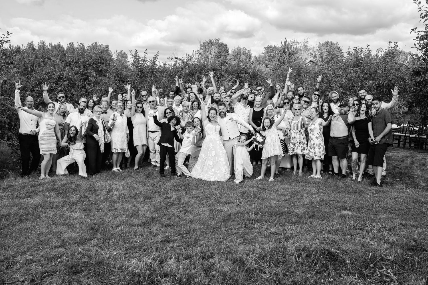 A joyful wedding group photo with the bride, groom, and guests celebrating outdoors in a lush green orchard.