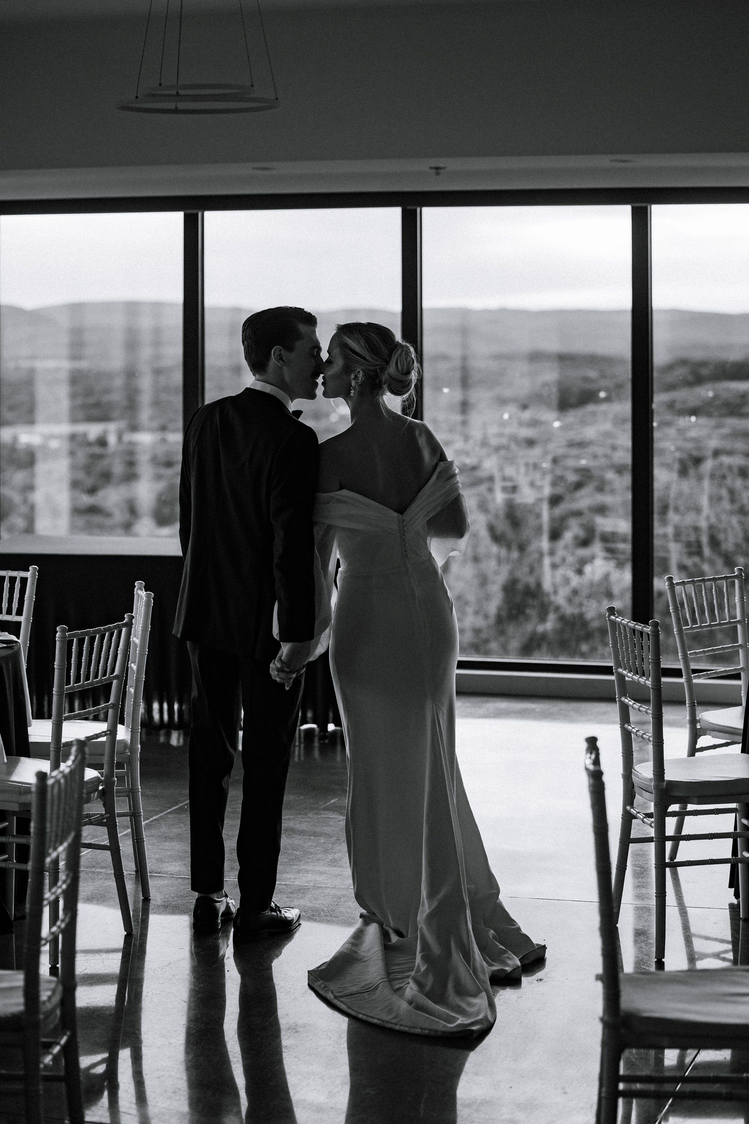 Black and white photo of bride and groom holding hands kissing by large venue windows.