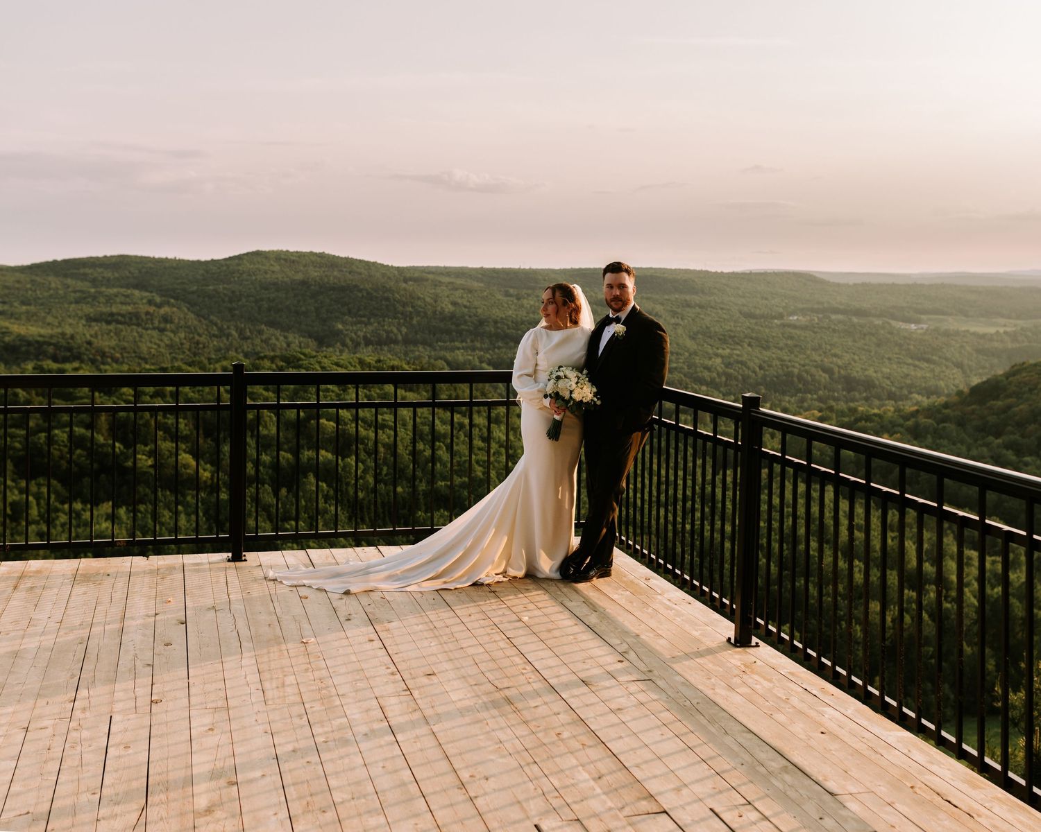 Bride and groom pose on scenic overlook deck with lush green hills and misty horizon at golden hour.