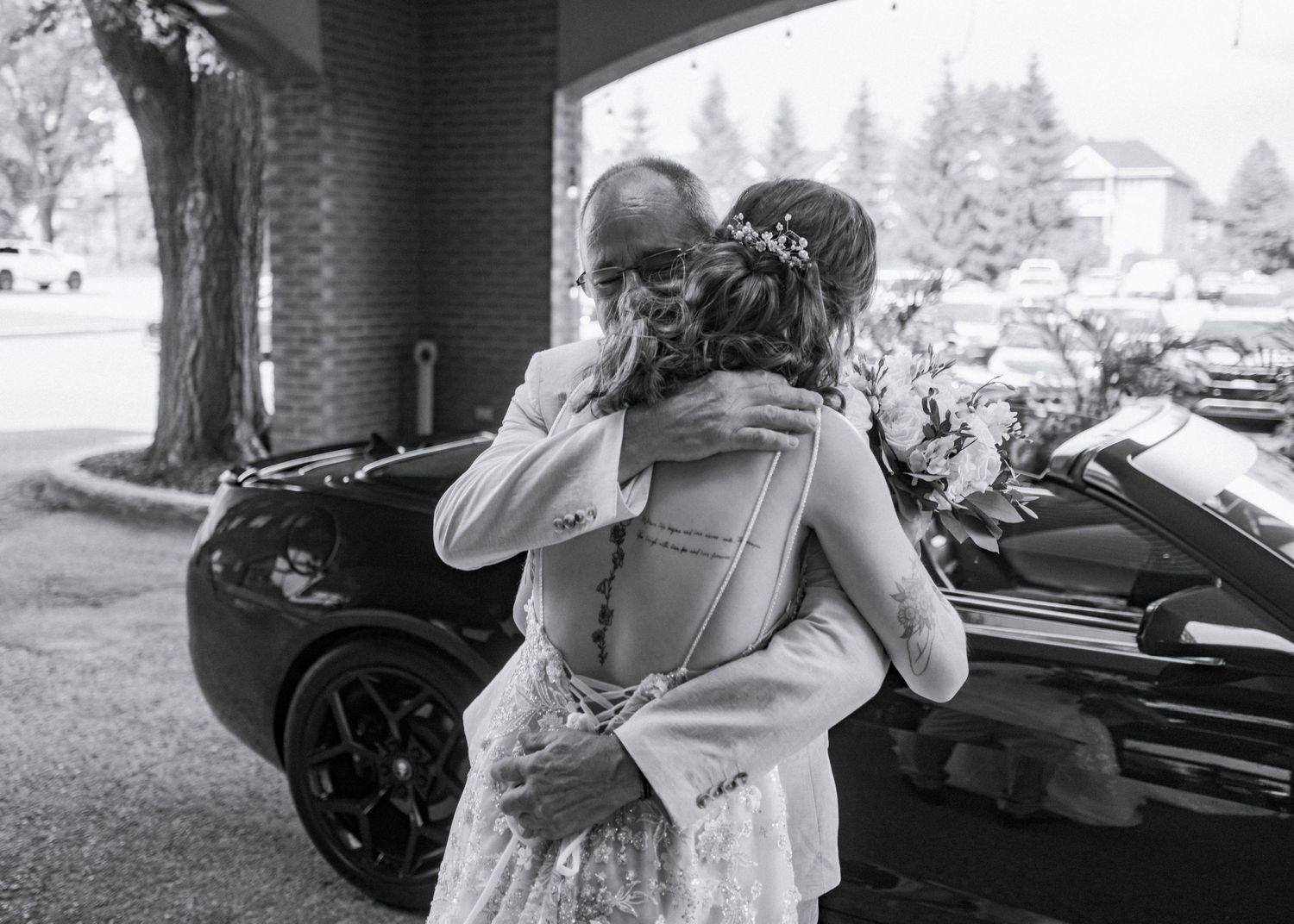 Black and white photo of a bride and elderly man sharing an emotional hug beside a luxury car outdoors.