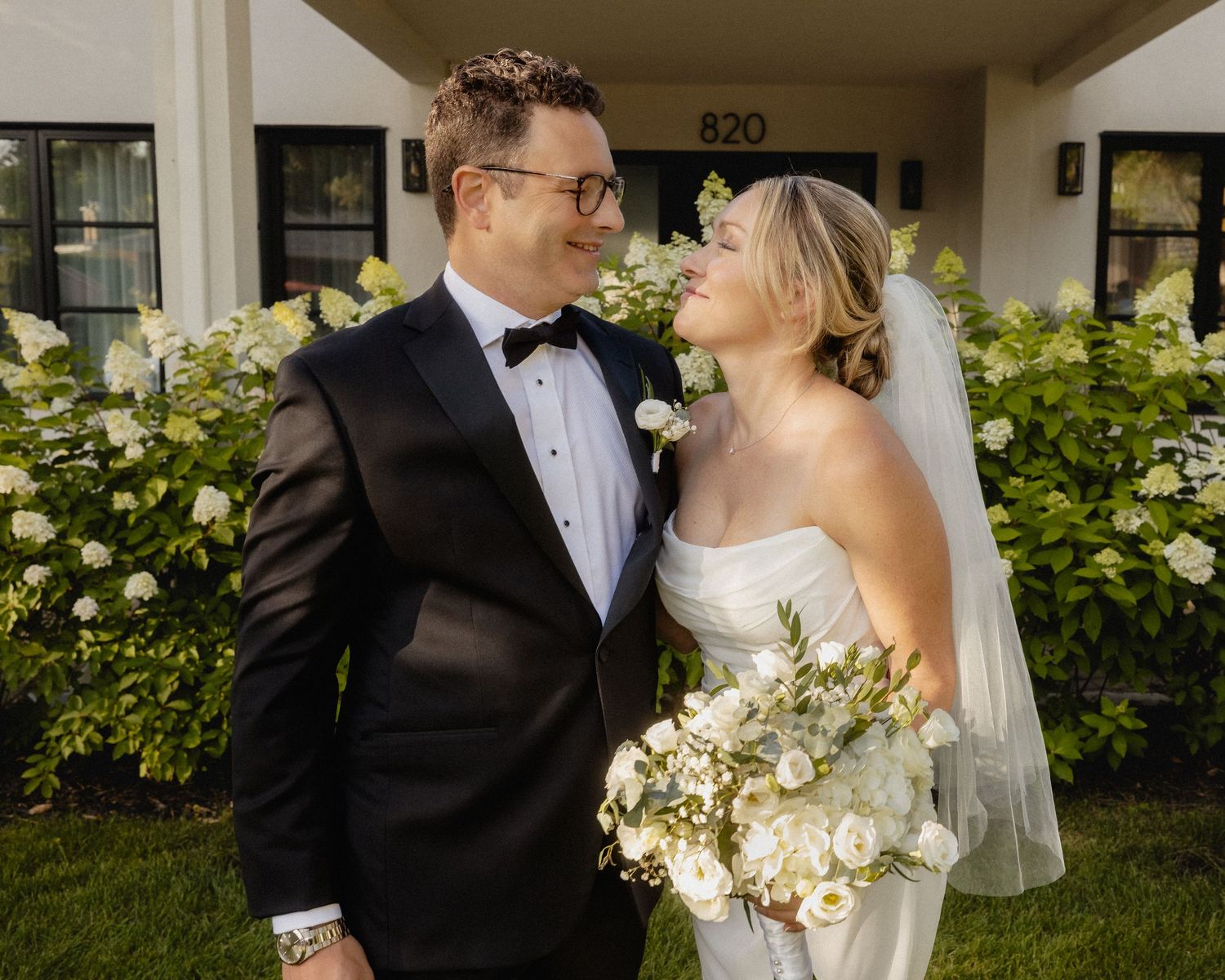 Bride and groom smiling at each other in front of white hydrangeas, she holds a white bouquet and wears a veil.