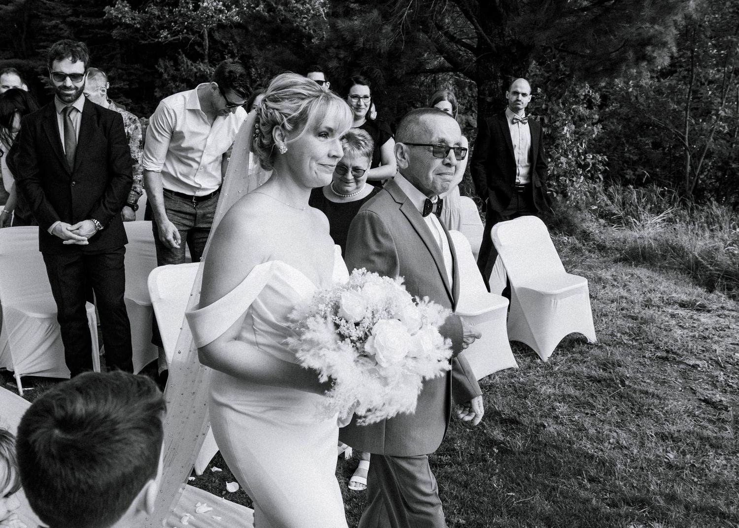 Bride in white gown walks down aisle with escort at outdoor wedding ceremony in black and white photo.