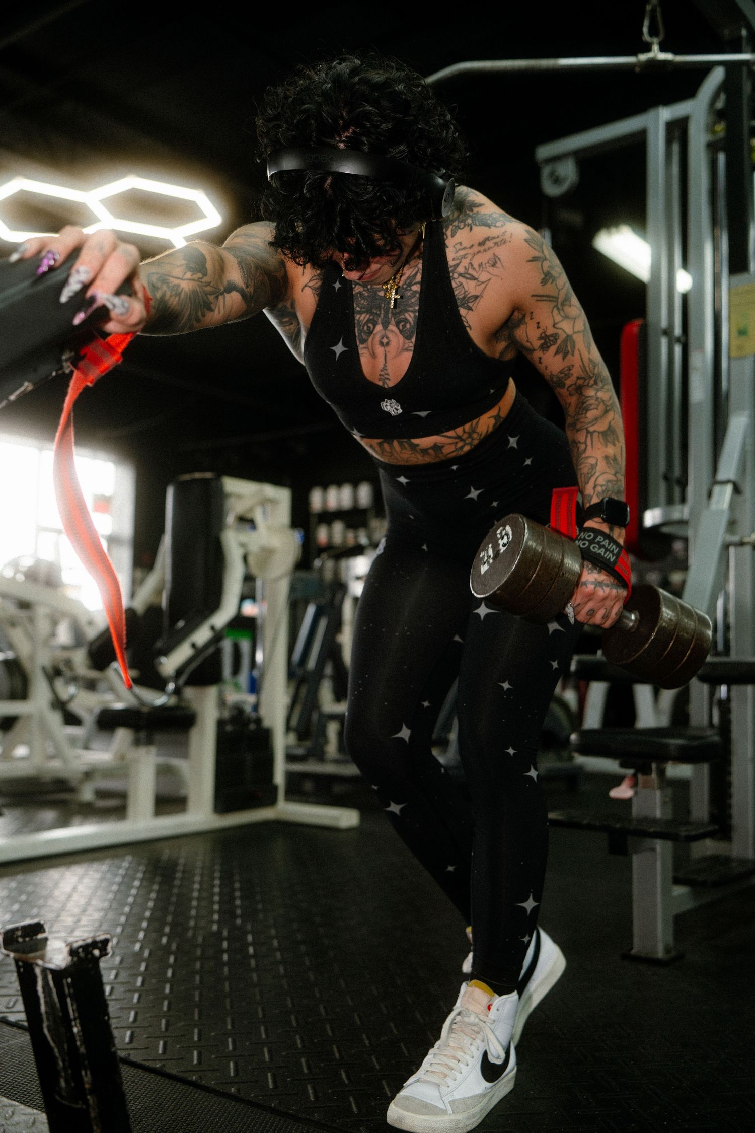 Heavily tattooed athlete in black workout gear lifting dumbbell intensely in a modern gym.